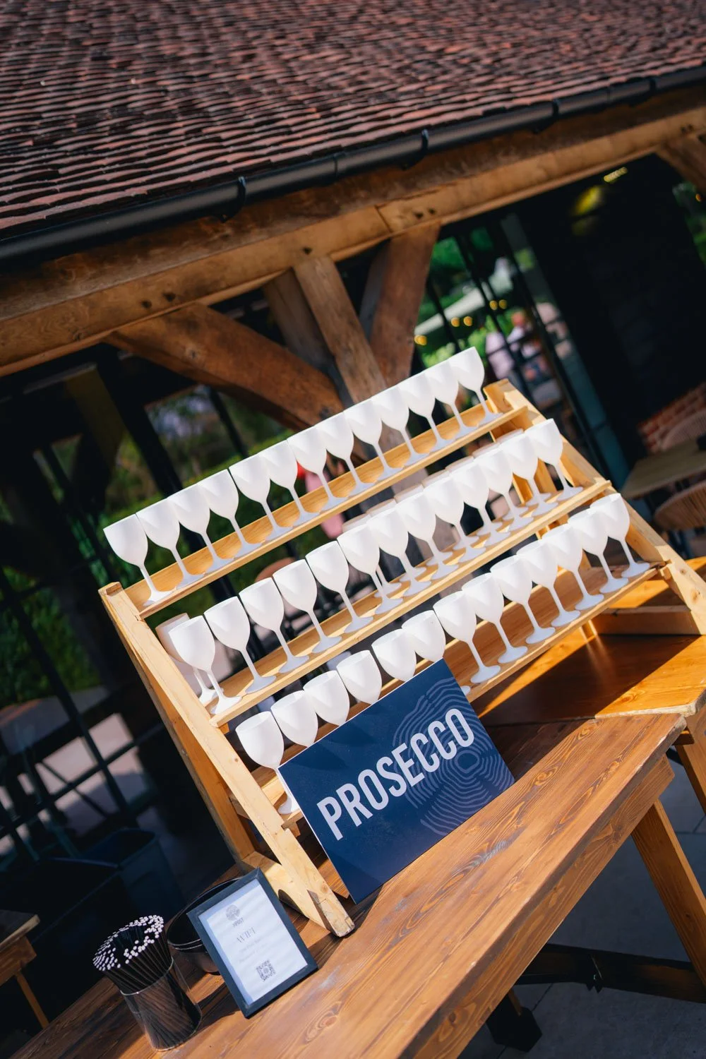 A photobooth with a sign that says Prosecco, featuring a stand with rows of white plastic glasses on wooden racks, set up outdoors on a wooden table under a roof with wooden beams.
