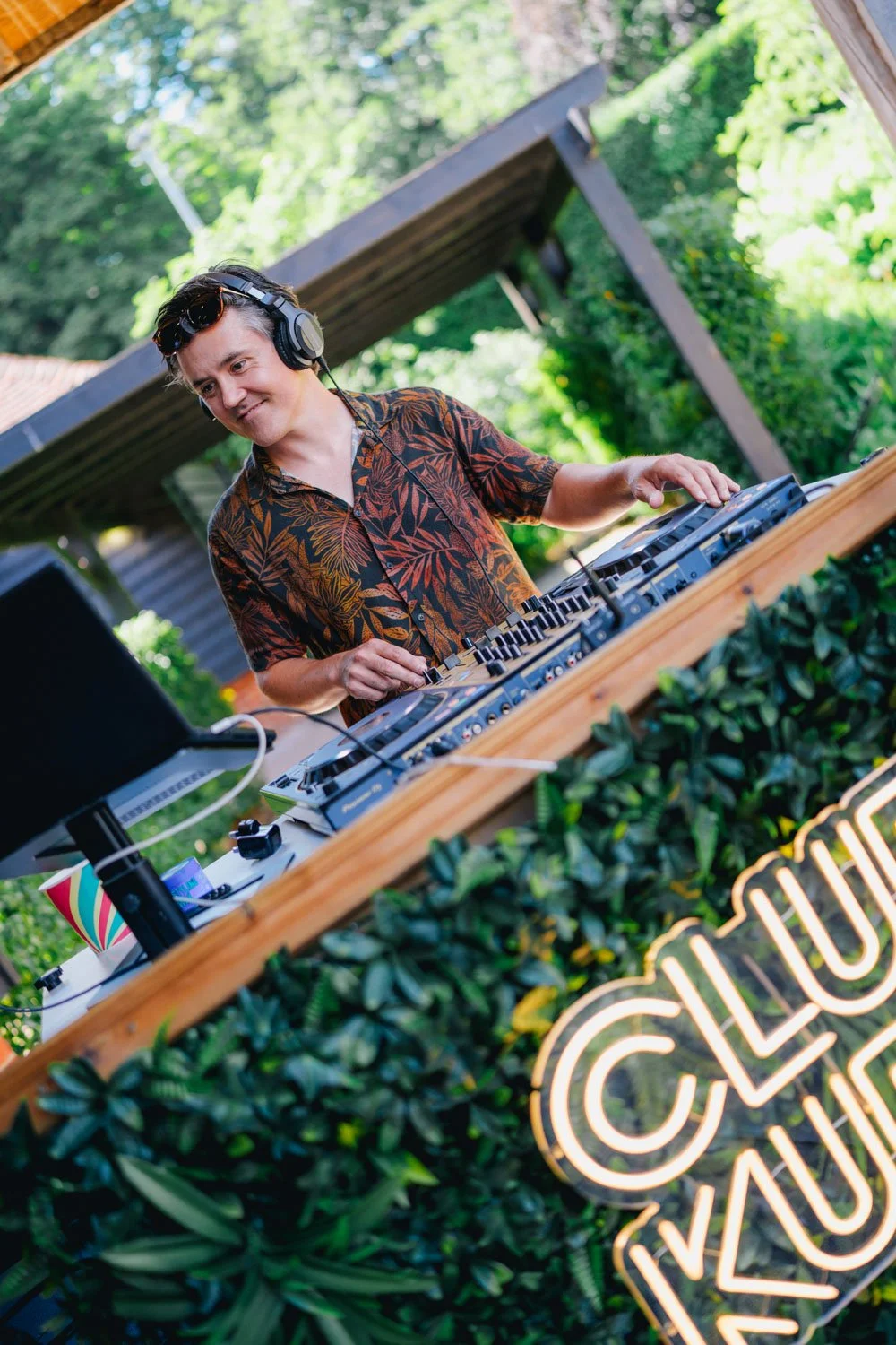 A man wearing a floral shirt and headphones DJing outdoors at a party, with greenery and a wooden structure in the background, and a neon sign that says 'Club Lounge' in front of the DJ setup.
