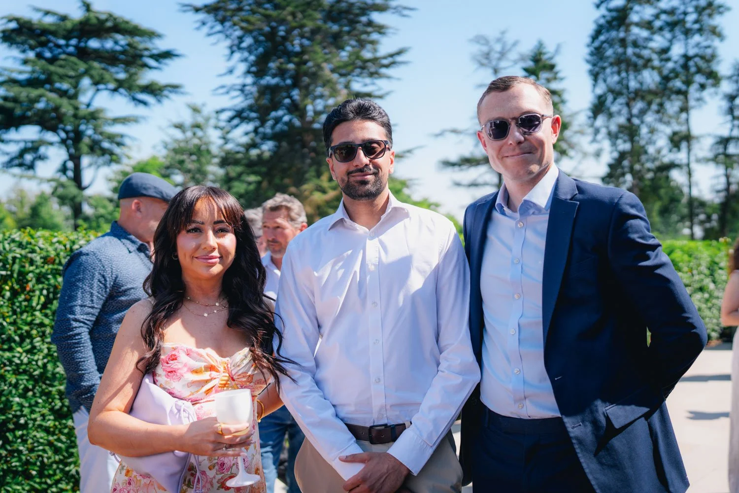 Three people standing outdoors at a sunny event, with two men wearing suits and sunglasses and a woman holding a wine glass, surrounded by other guests and greenery.