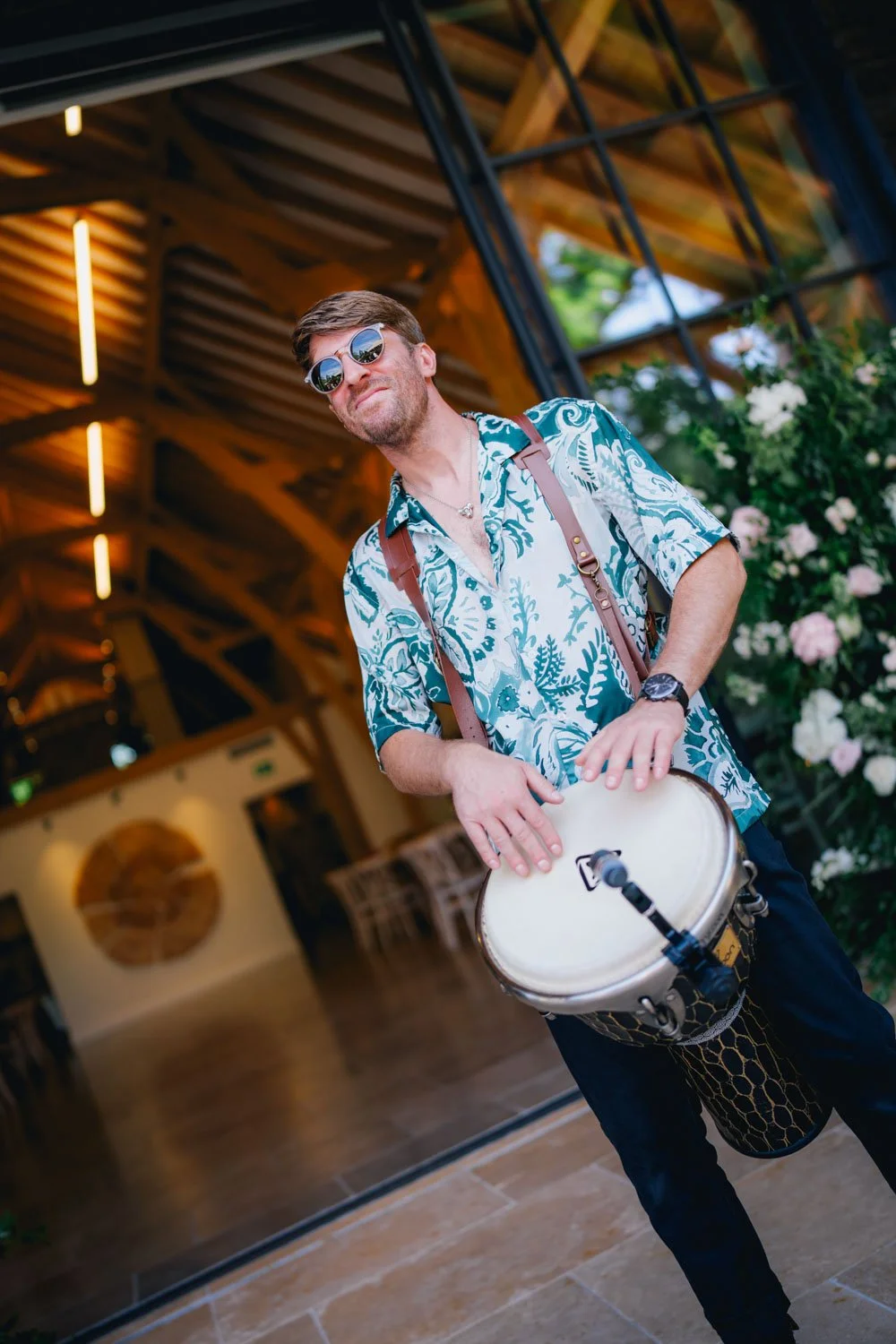 A man wearing sunglasses and a floral shirt playing a drum inside a wooden building with plants and flowers outside.