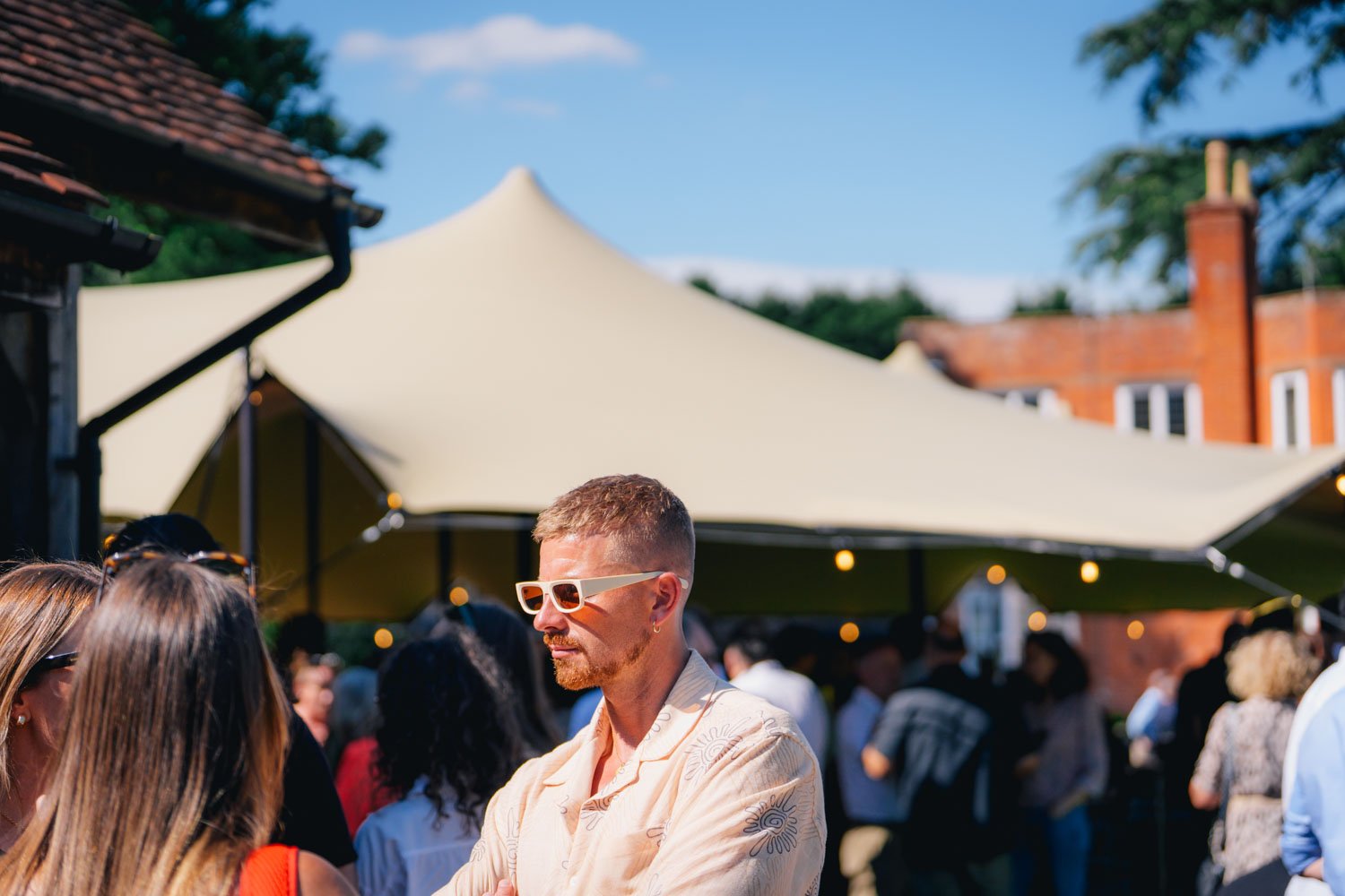 People socializing at an outdoor party under a large beige canopy tent with string lights, in a neighborhood with trees and brick houses.
