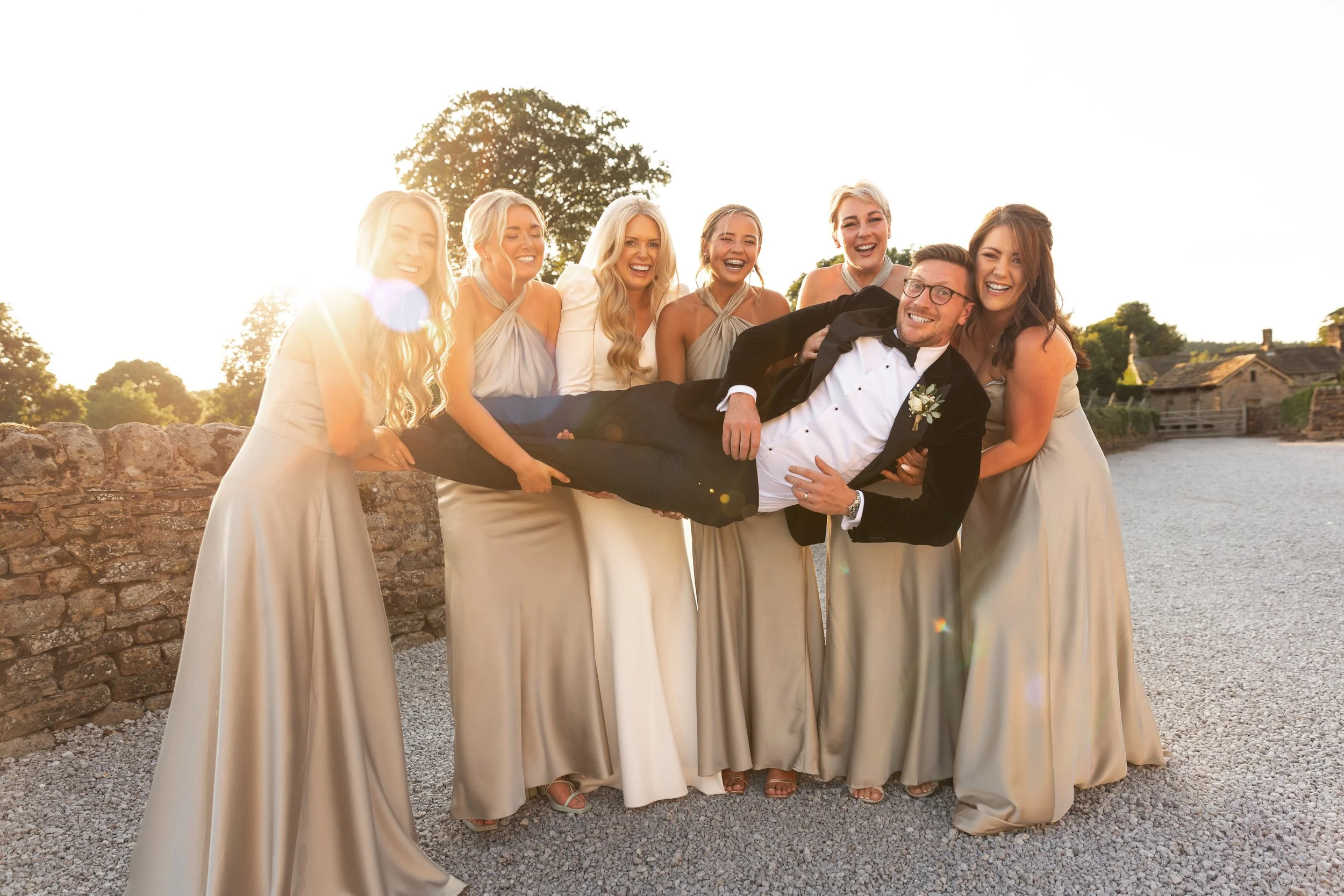 Group of seven women and one man, dressed in formal attire, outdoors at sunset, with the man being held horizontally by the women, all smiling and laughing, near a stone wall and a water body.