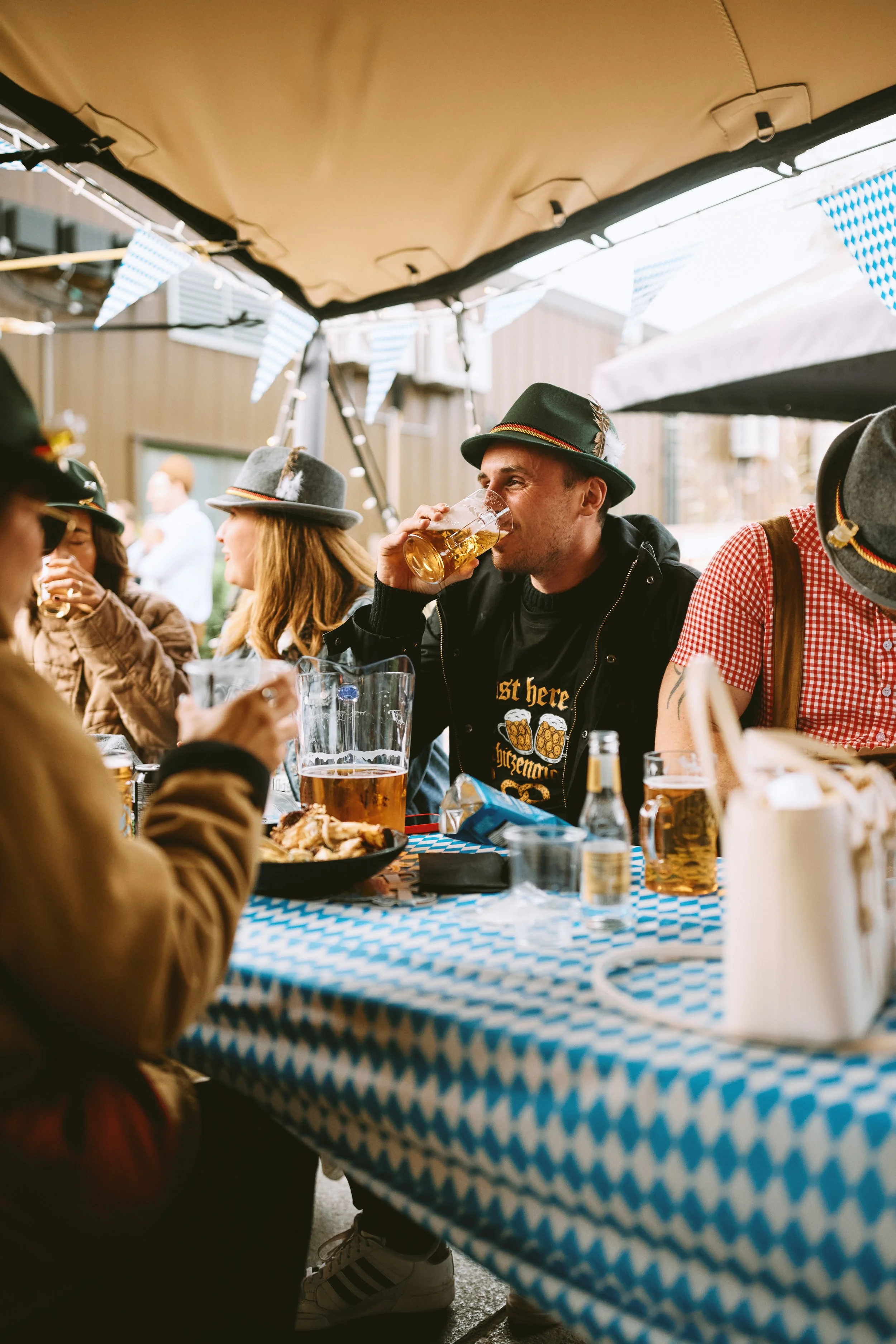 People celebrating at a festive outdoor gathering, wearing traditional German attire, with beer in hand, under blue and white decorations.