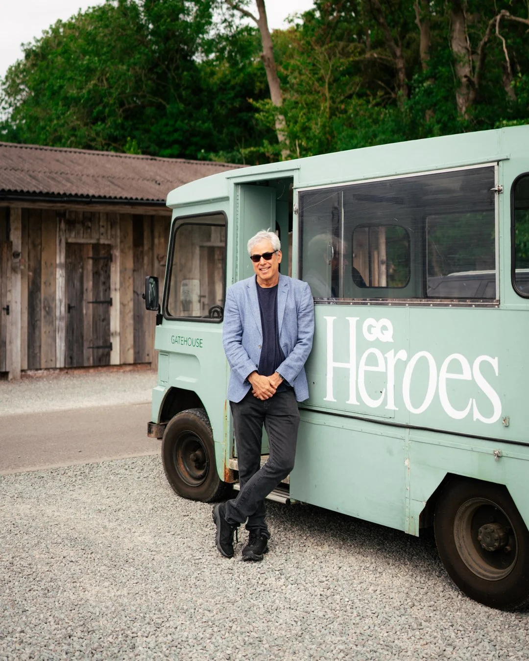 A man in sunglasses and a blazer leaning against a light green food truck with 'GQ Heroes' logo, parked on gravel near a wooden building and green trees.