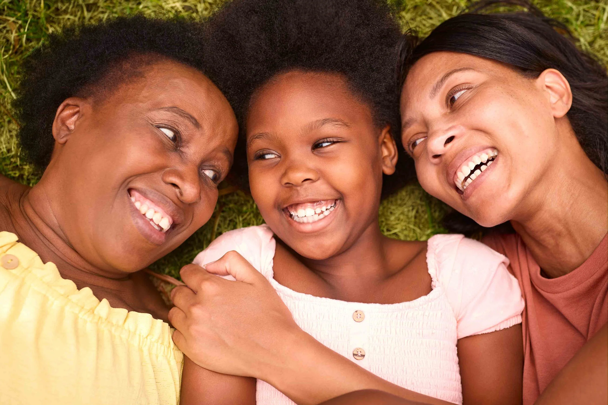 Three women and a young girl lying on grass, smiling and looking at each other.