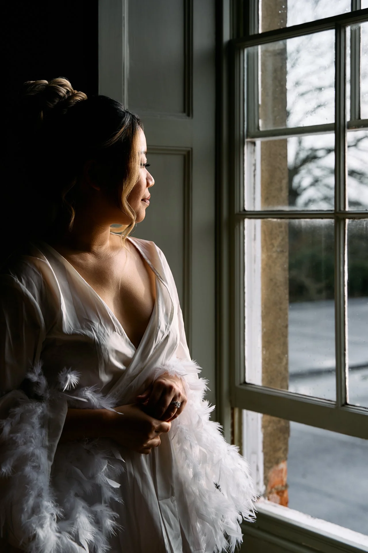 A woman with styled hair and wearing a silky white dress with feathers is looking out a window on a cloudy day, with her hands clasped in front of her.
