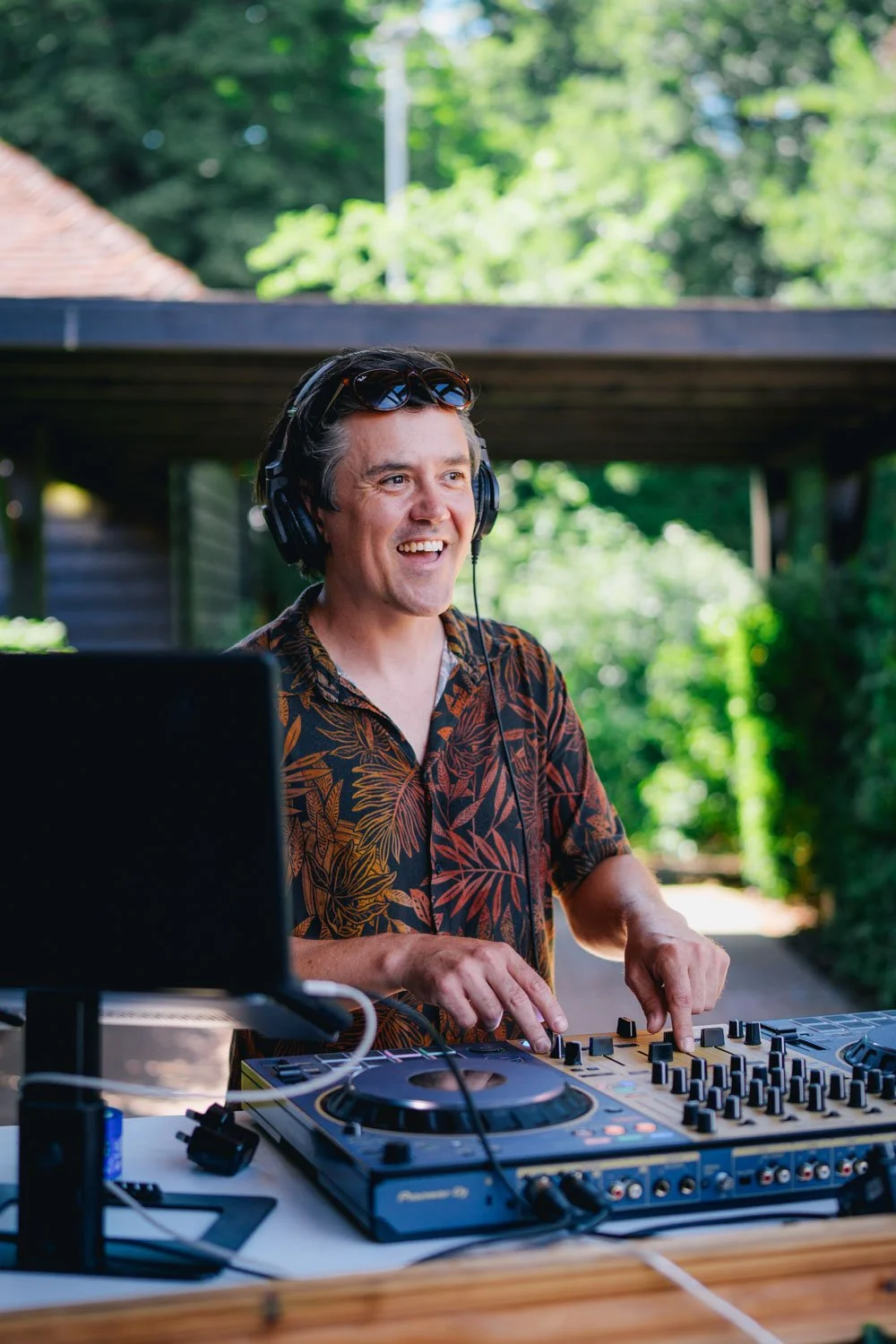 A man wearing headphones, a patterned shirt, and sunglasses on his head, smiling while DJing outdoors.