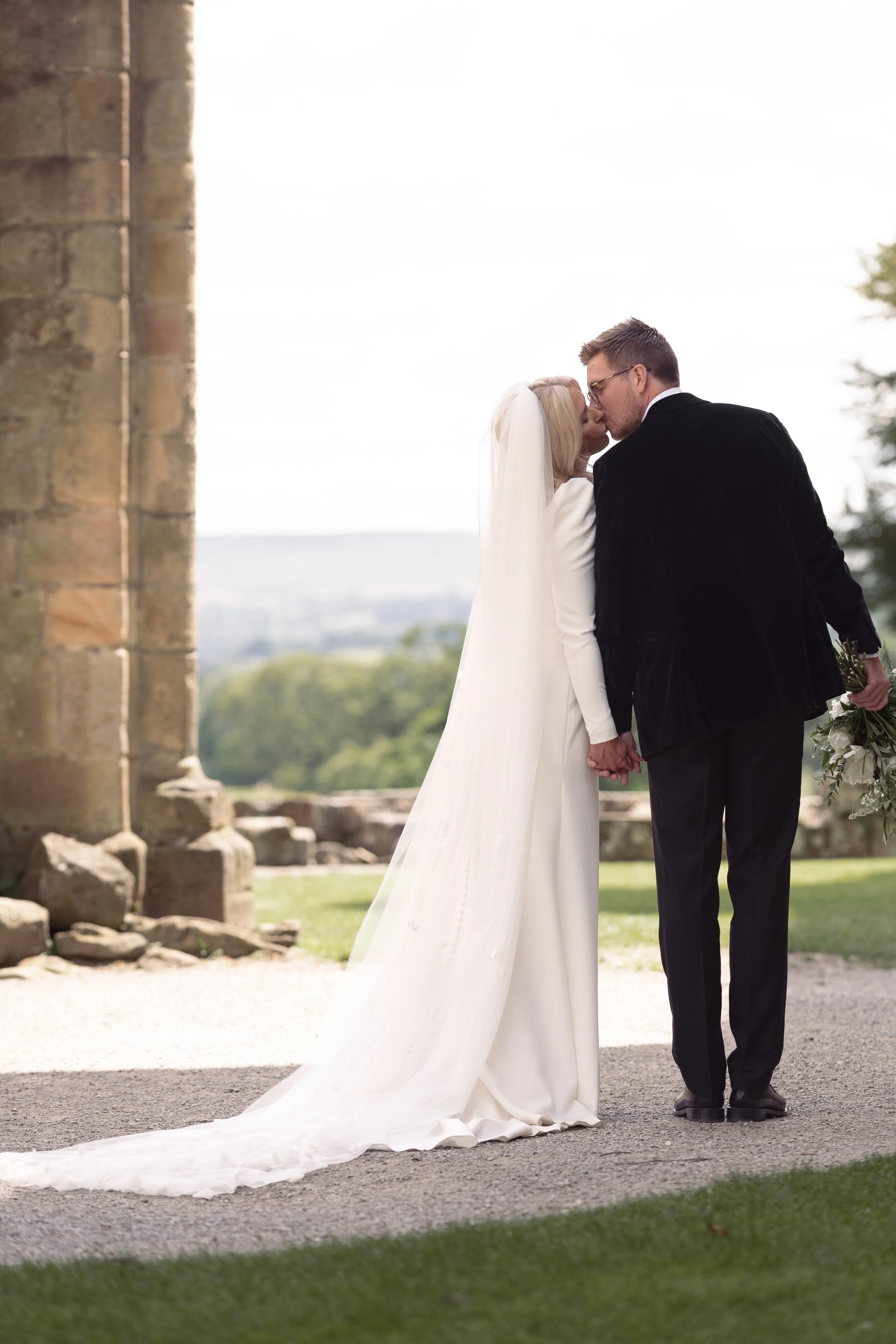 A bride and groom kissing outdoors during their wedding, holding hands, with the bride in a white dress and veil, and the groom in black attire, near a stone structure.