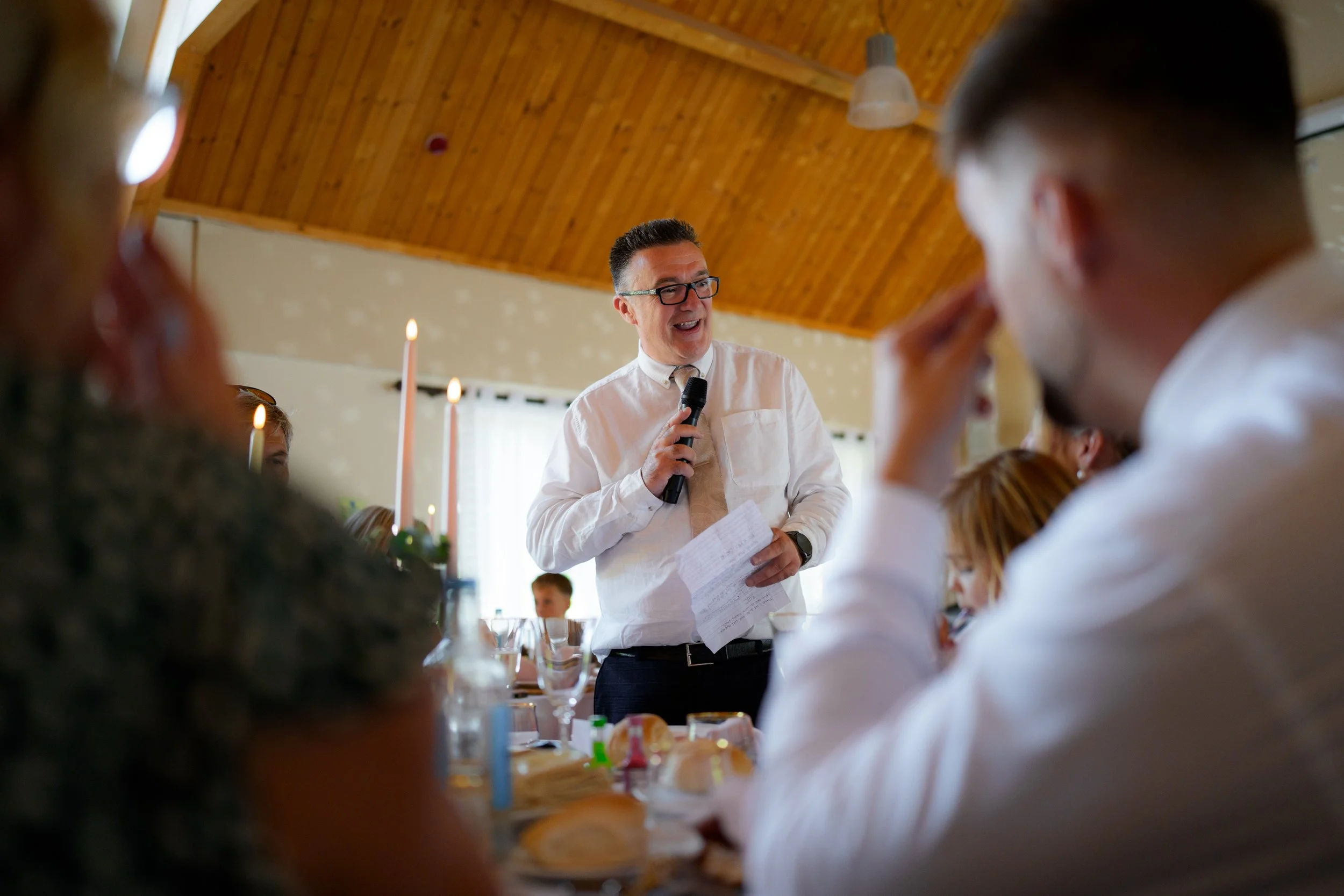 A man in a white shirt and glasses holding a microphone and paper, speaking to a group in a room with wooden ceiling, surrounded by people seated at a table with lit candles and glasses.