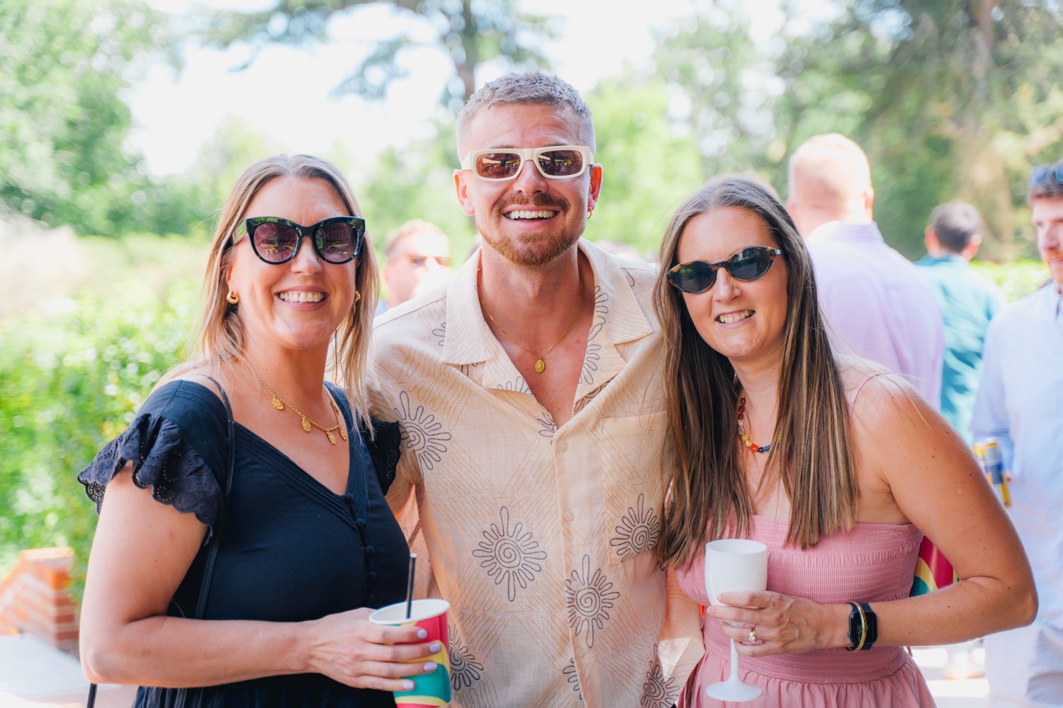 Three people wearing sunglasses smiling outdoors at a daytime gathering, with trees in the background, holding drinks.