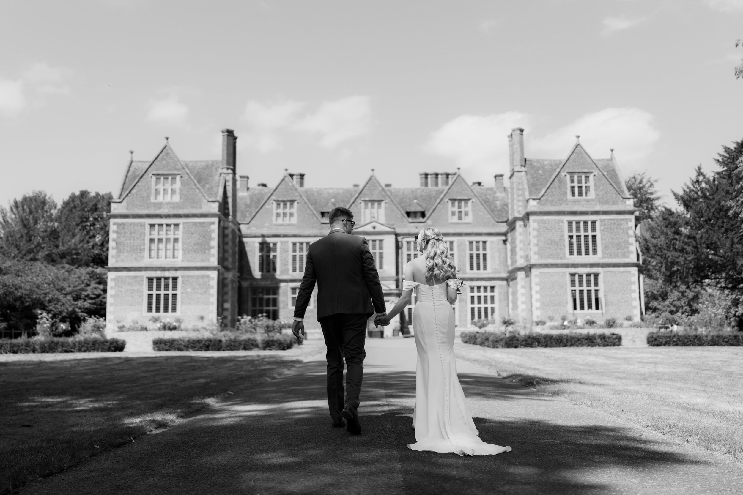 Black and white photo of a bride and groom walking hand in hand on a pathway in front of a large, historic mansion surrounded by greenery.