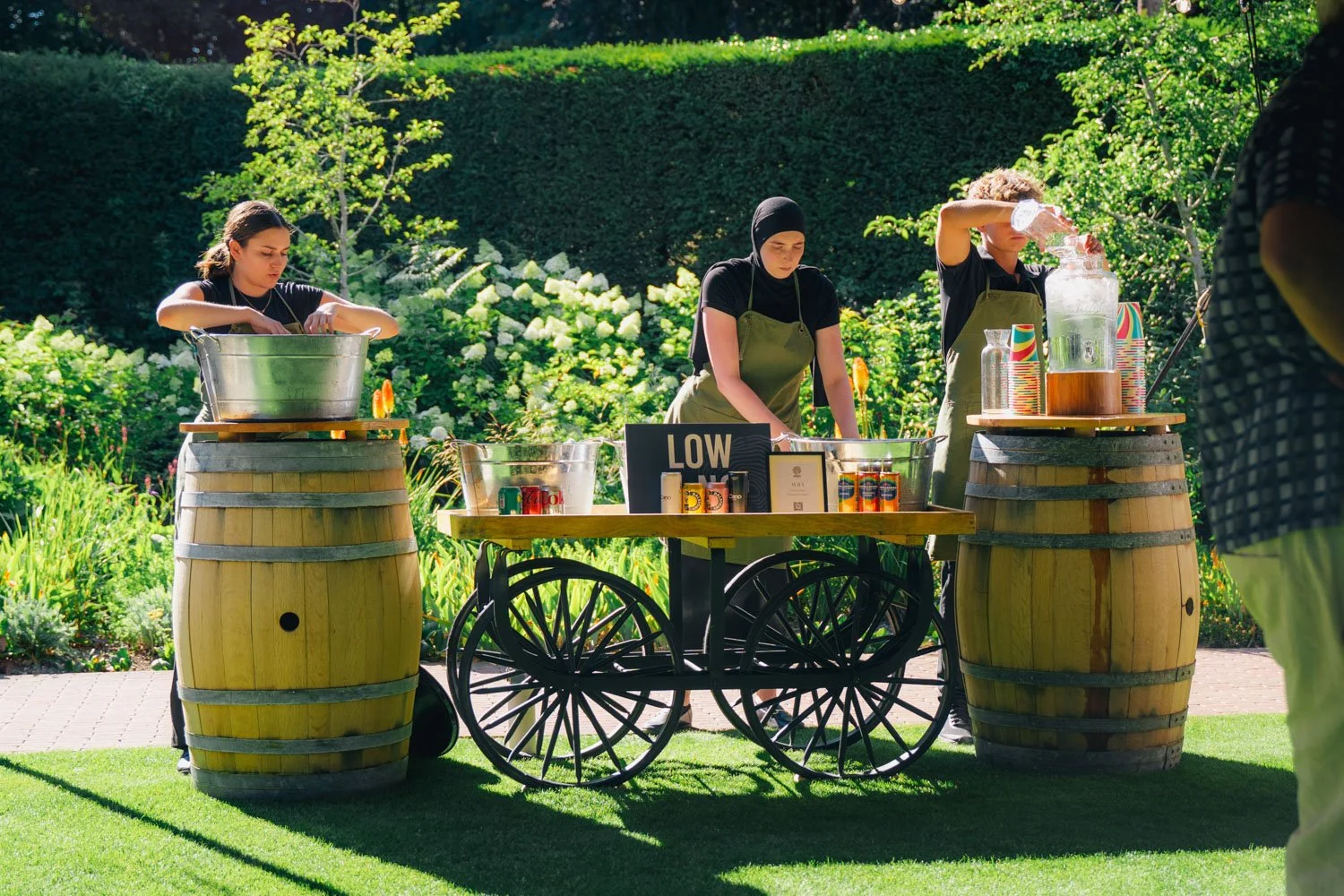 Three women working at a lemonade stand made of barrels and a cart with a sign that says "LOW" in a lush, green garden.