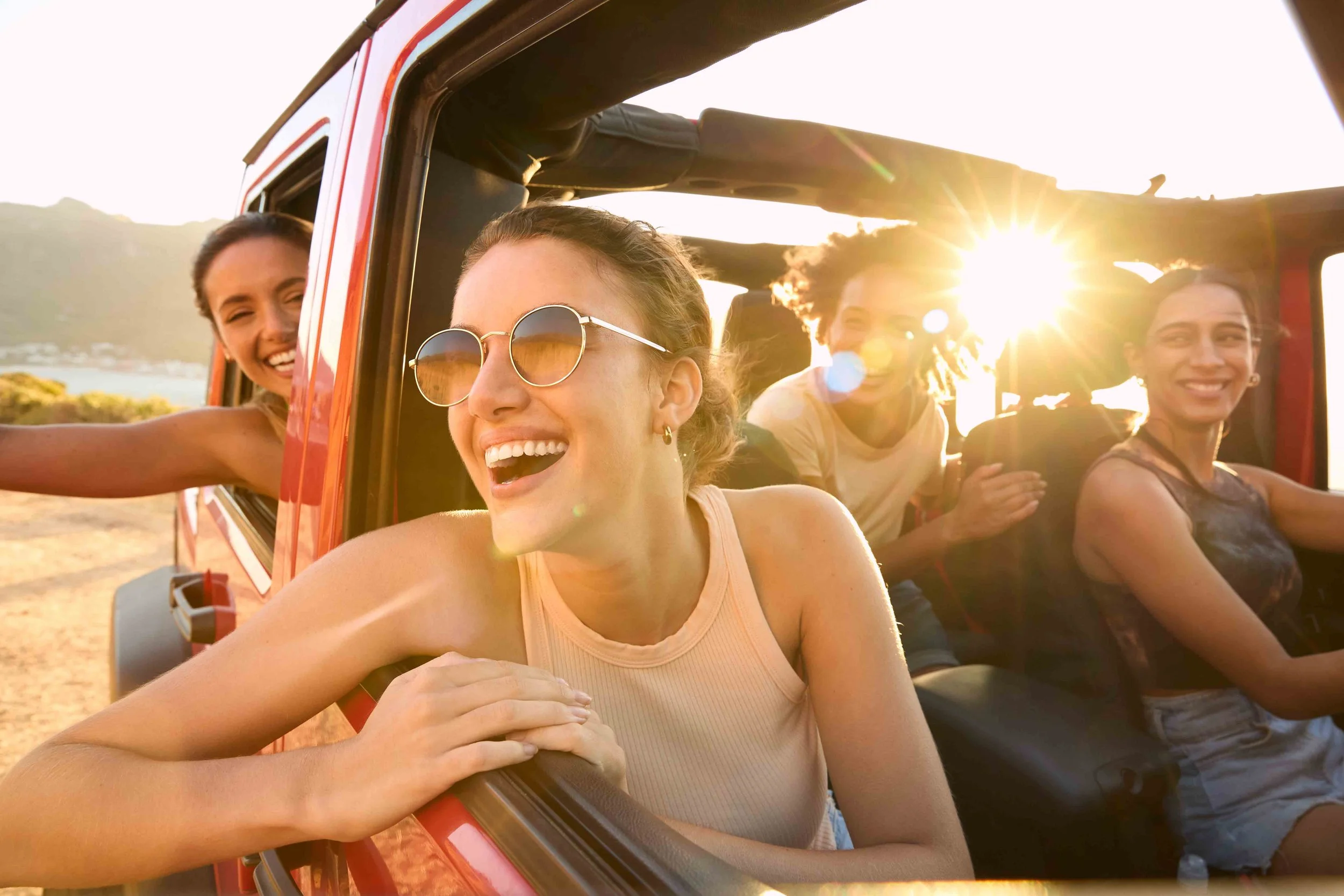 Four young women enjoying a ride in a red off-road vehicle with open windows during sunset, smiling and relaxing in a scenic outdoor location.