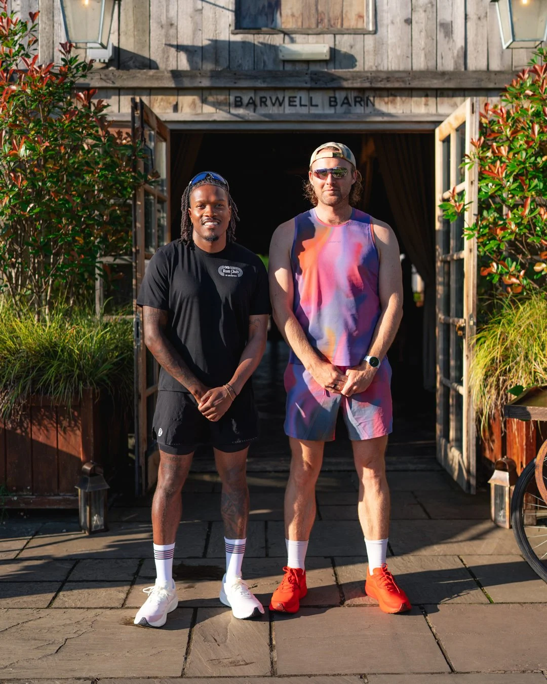 Two men standing outdoors in front of a building with a sign that reads 'Barwell Barn'. The man on the left has dreadlocks, tattoos, and is dressed in a black sports shirt and shorts, wearing white sneakers and socks. The man on the right has shoulde