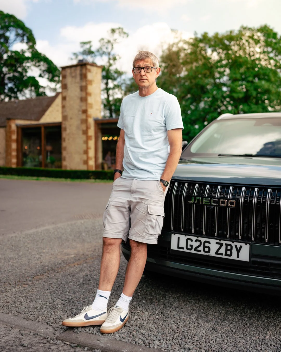 A man in shorts and a white t-shirt standing with hands in pockets next to a black car with a license plate LG26 ZBY and grille with the brand name 'JAECOO' in a suburban neighborhood with houses and trees in the background.