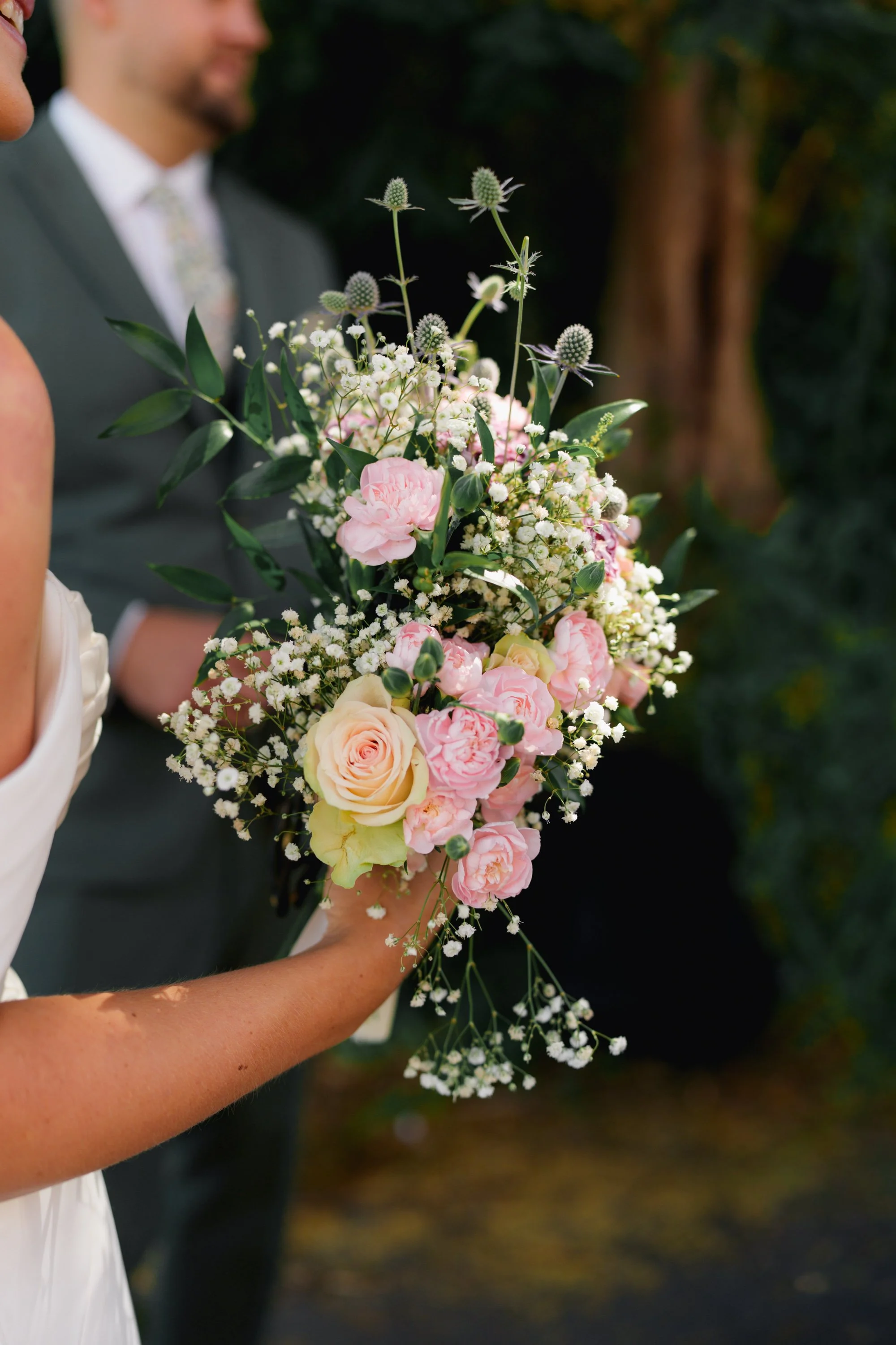 A person holding a large bouquet of pink, white, and cream roses with greenery and small white flowers, in an outdoor setting with a blurred background.