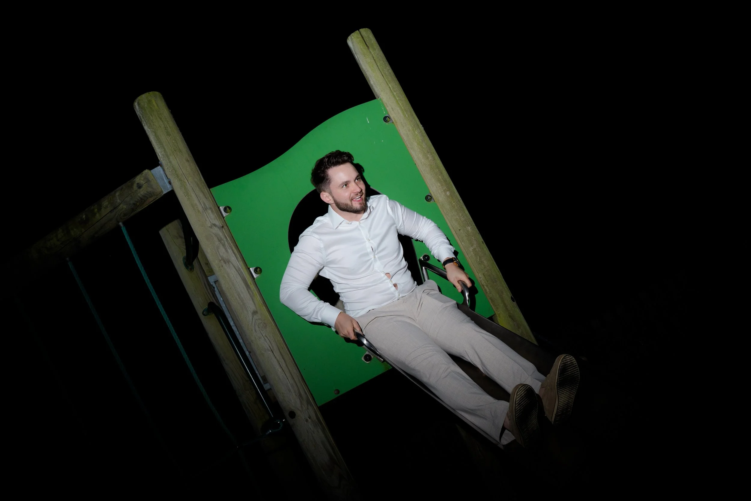 A man in a white shirt and beige pants is sitting on a green slide at night.