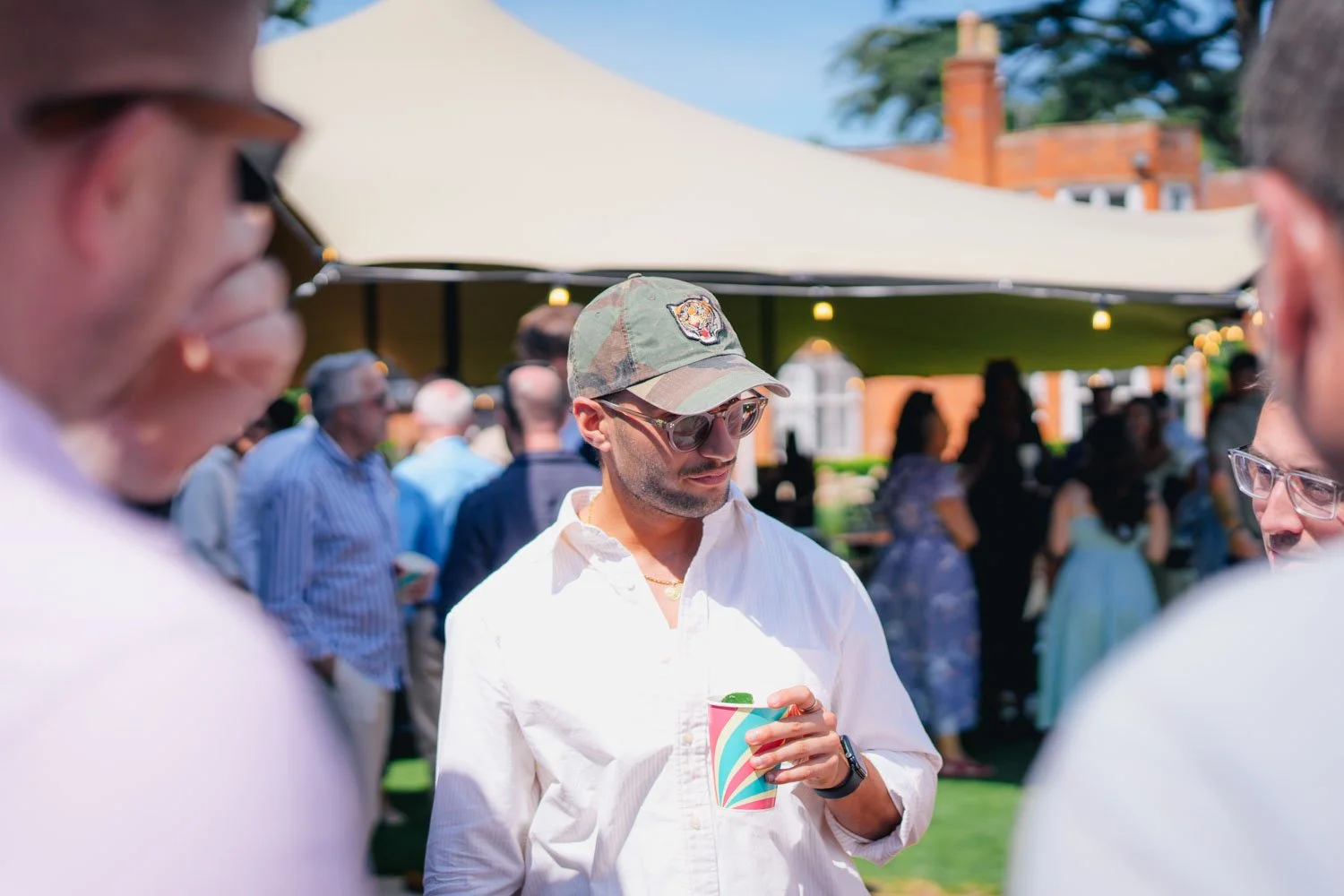 A man wearing a camouflage baseball cap, sunglasses, and a white shirt, holding a colorful cup at an outdoor social gathering on a sunny day.