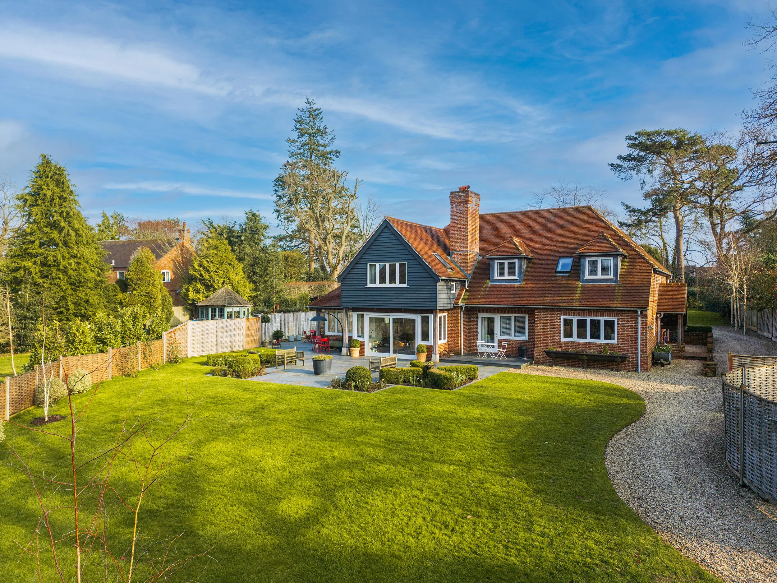 Large brick house with a red-tiled roof and a chimney, surrounded by a well-maintained lawn and trees, under a blue sky with scattered clouds.