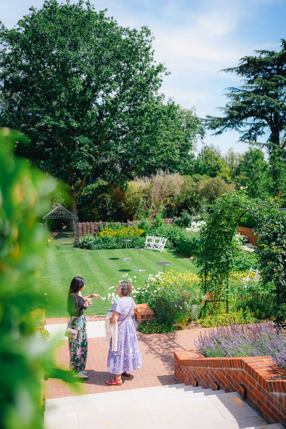 Two women talking in a lush garden with green trees, flowering plants, and a manicured lawn on a sunny day.