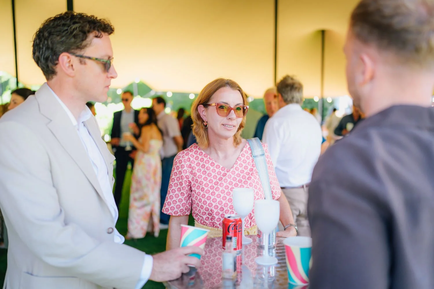 People mingling at an outdoor event under a tent, with a woman in pink patterned dress and sunglasses talking to two men, one in a cream blazer and the other in a dark jacket, with drinks on the table in front of them.