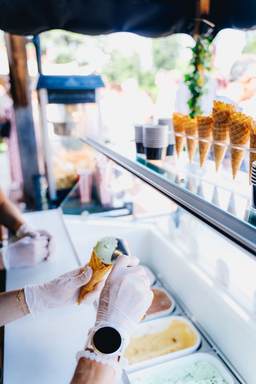 Person scooping green ice cream into a cone at an outdoor ice cream stand with waffle cones and cups of ice cream on display.
