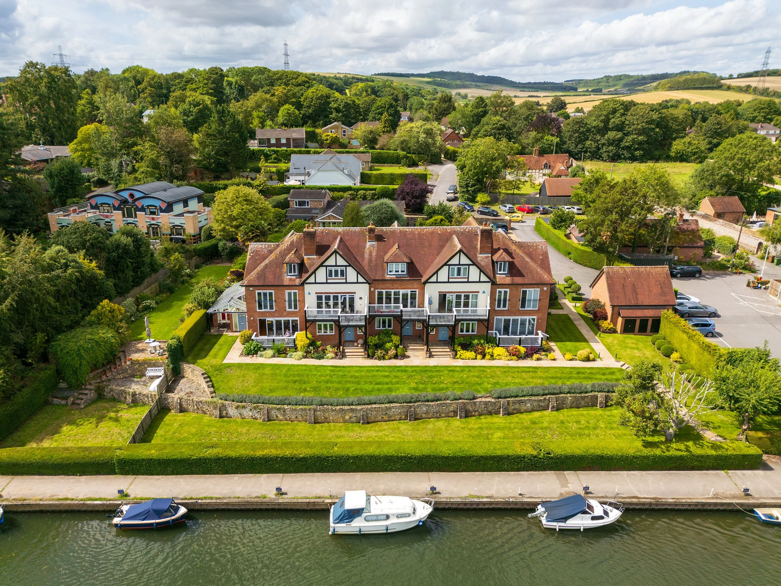 Aerial view of a large brick house with white framing and multiple gables, surrounded by green lawns and gardens, located along a river with boats docked at the shoreline. Neighborhood houses and trees are visible in the background.