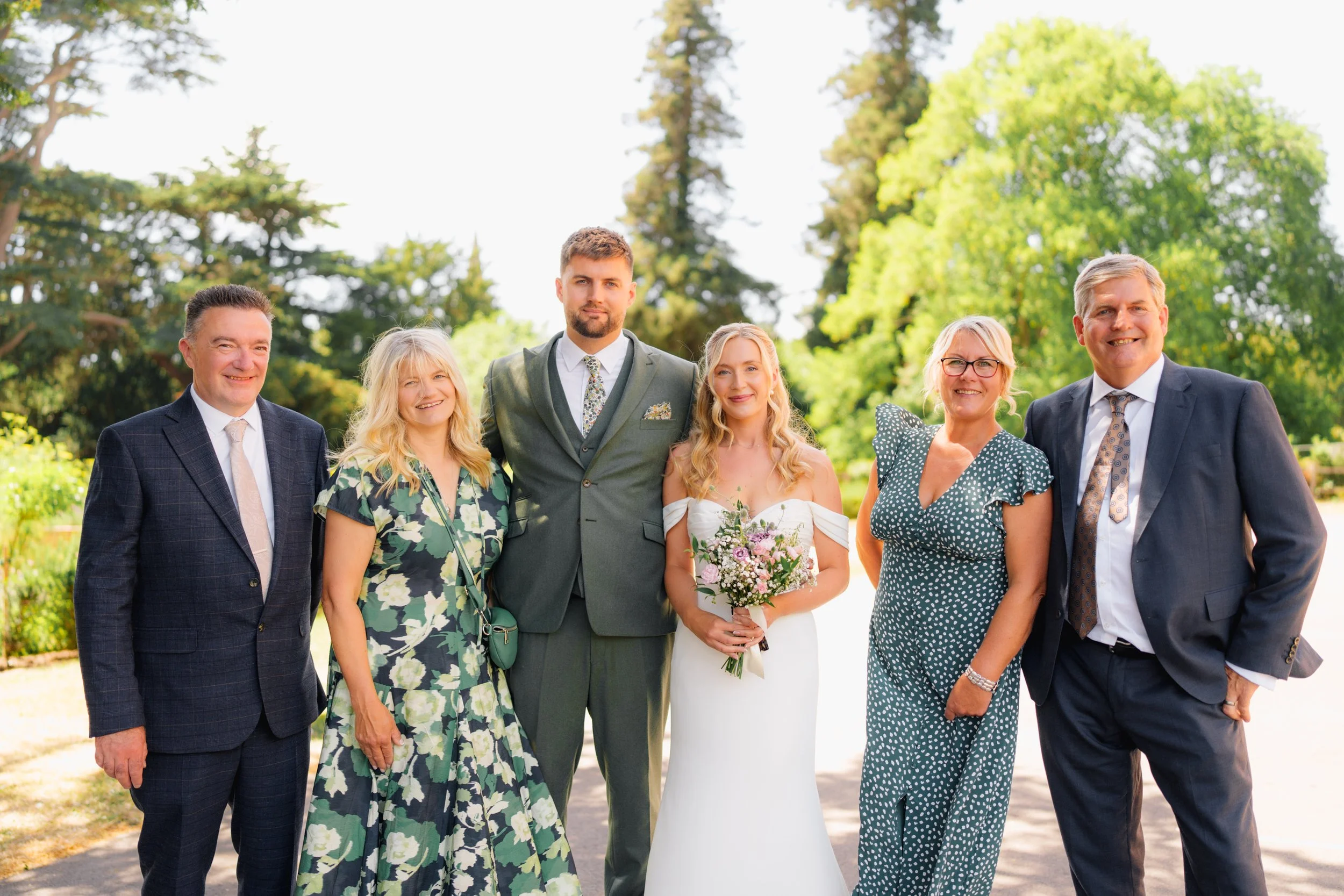 Group of seven people dressed formally, standing outdoors in front of green trees, smiling for a photo.