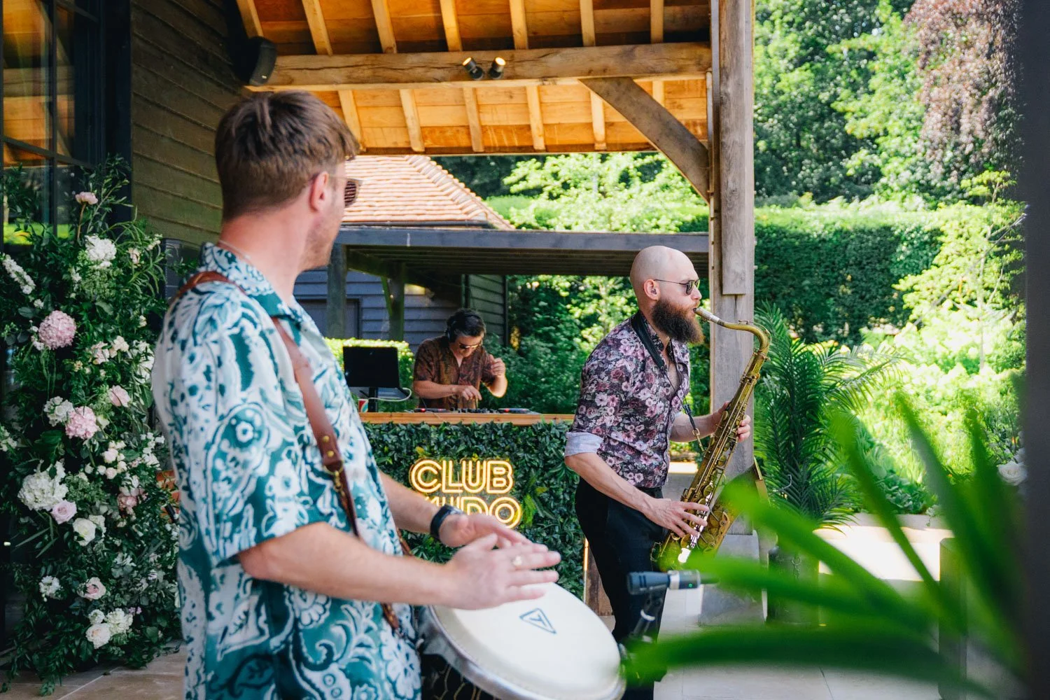 Three musicians perform outdoors at a lush garden venue, with one playing bongos, a second on a saxophone, and a DJ in the background behind a decorated table.