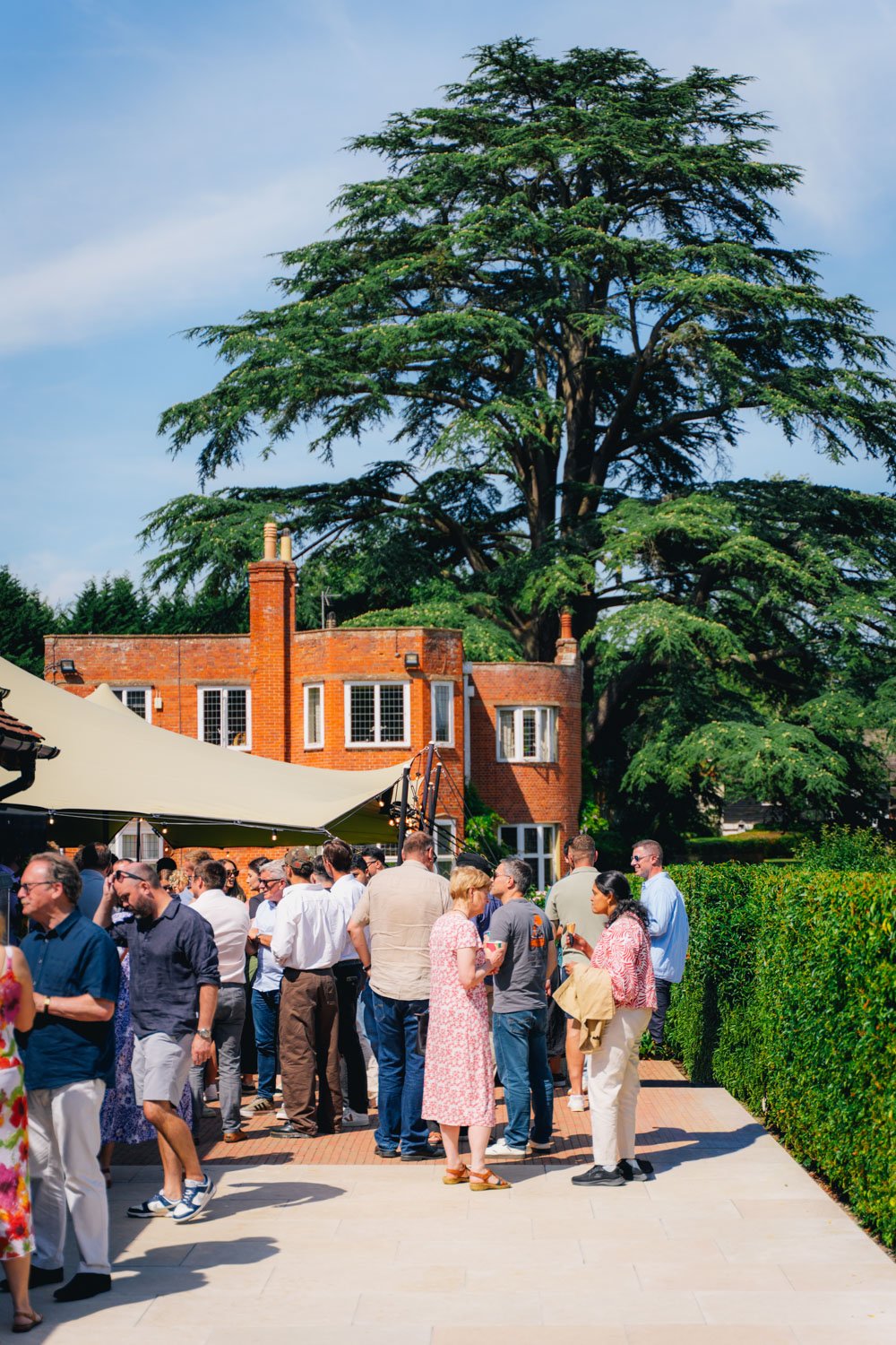 People gathered outdoors at a social event in a backyard, with a large green tree and a red brick house in the background.
