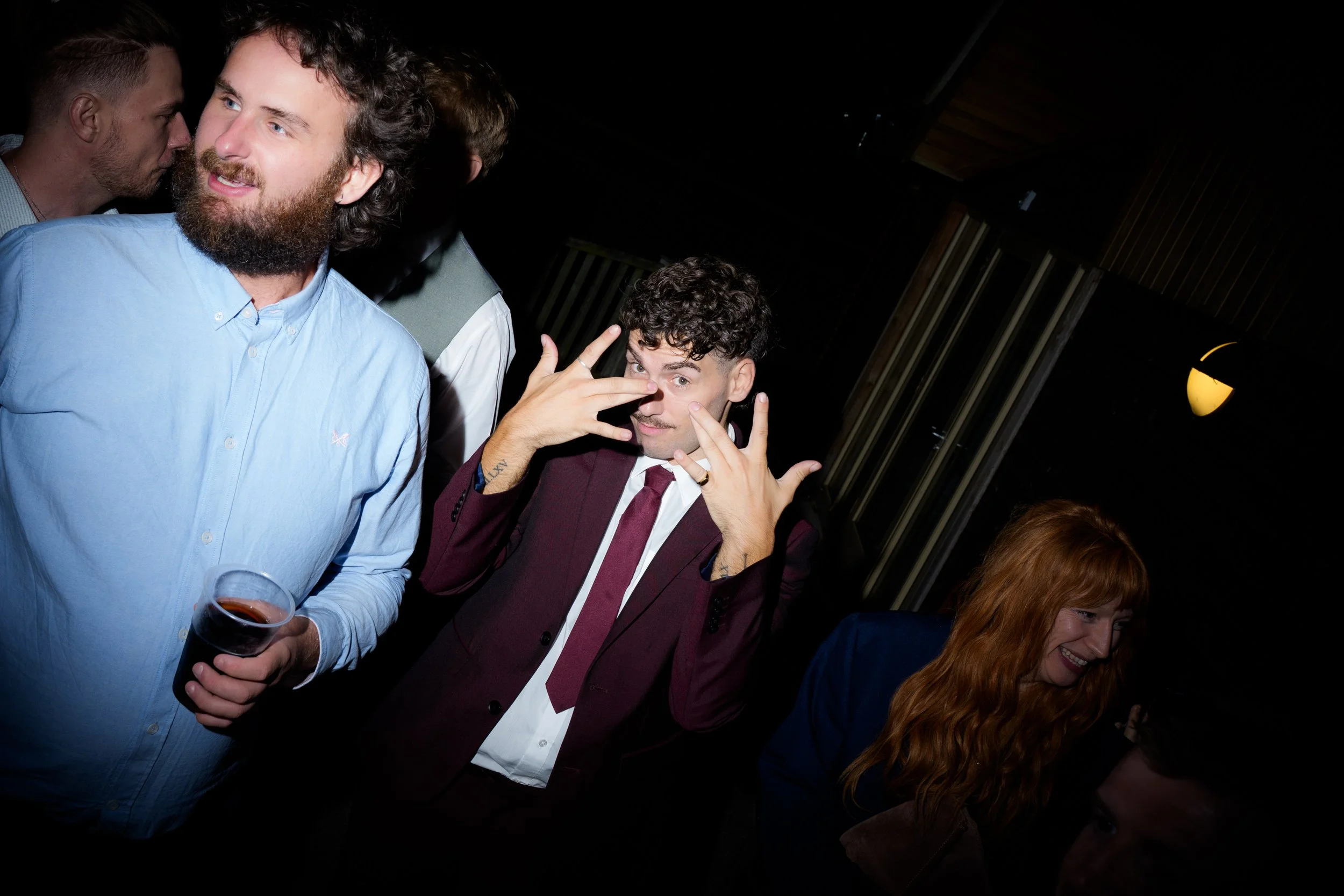 Group of people at a social gathering, with one man in a light blue shirt holding a drink, and another man in a burgundy blazer making a playful gesture near his face, in a dark indoor setting.