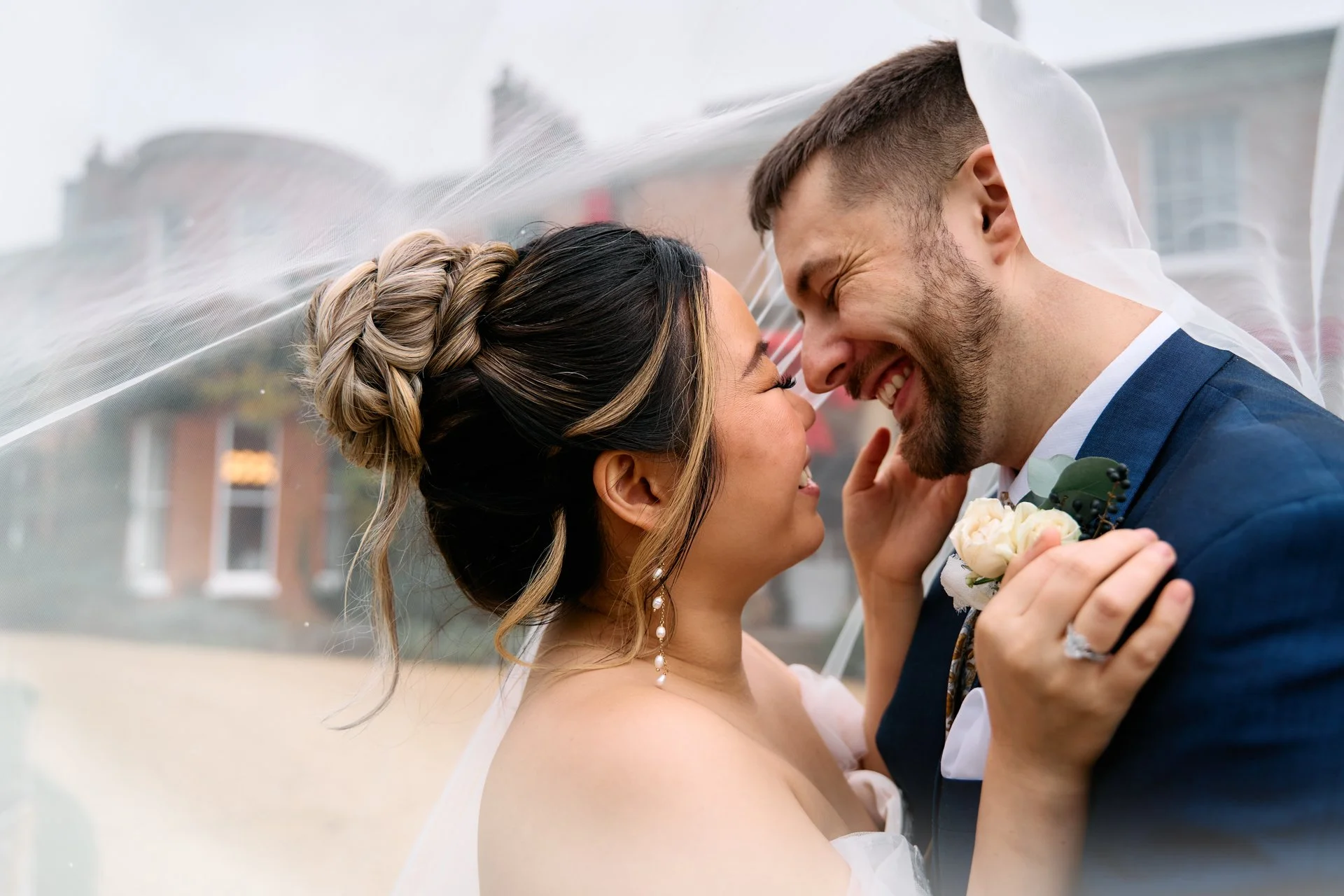 A bride and groom smiling and touching noses under a veil during their wedding outside, with a building in the background.