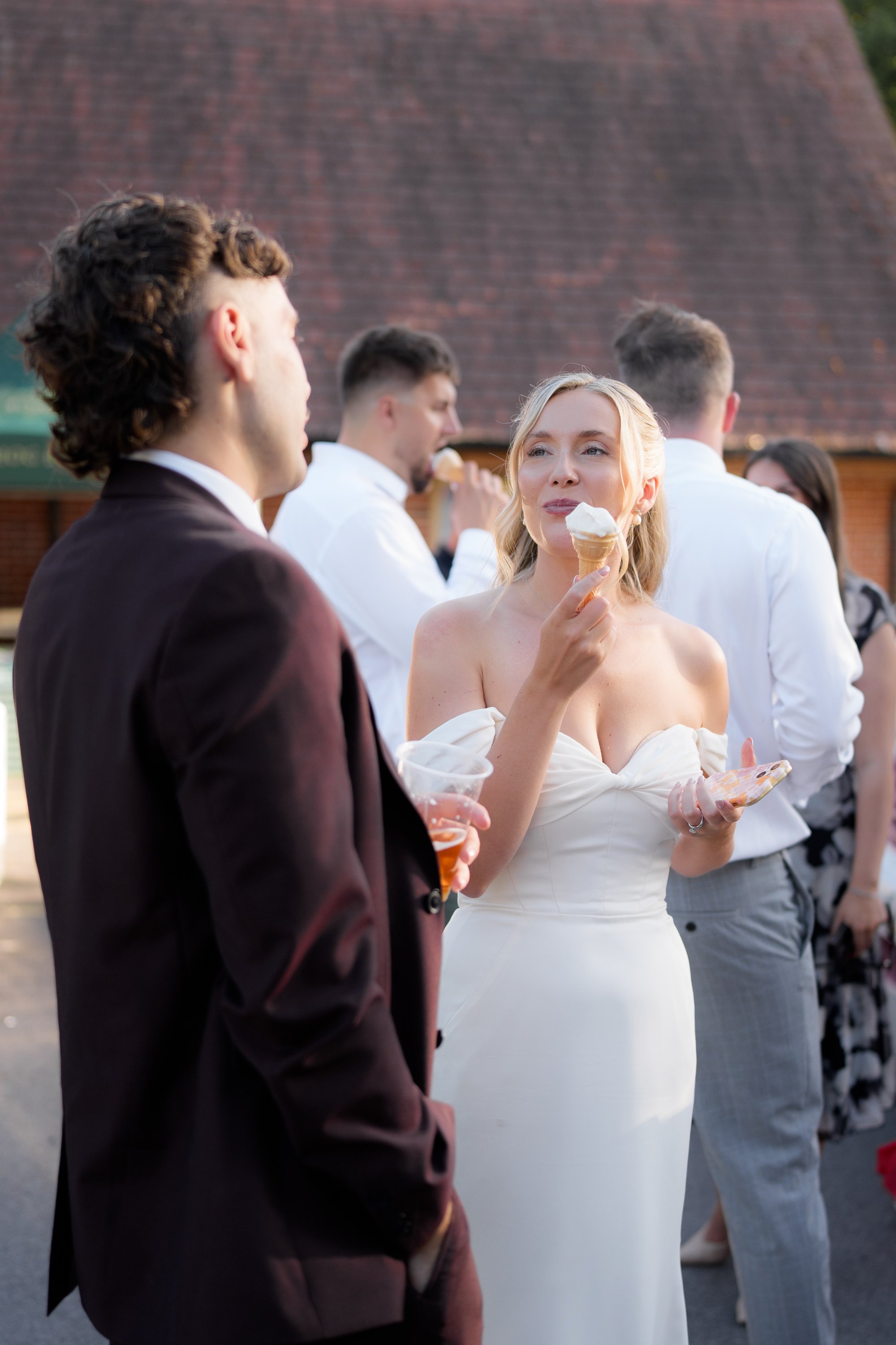 Young woman in off-shoulder white dress holding an ice cream cone, standing outdoors among people dressed formally, possibly at an event.