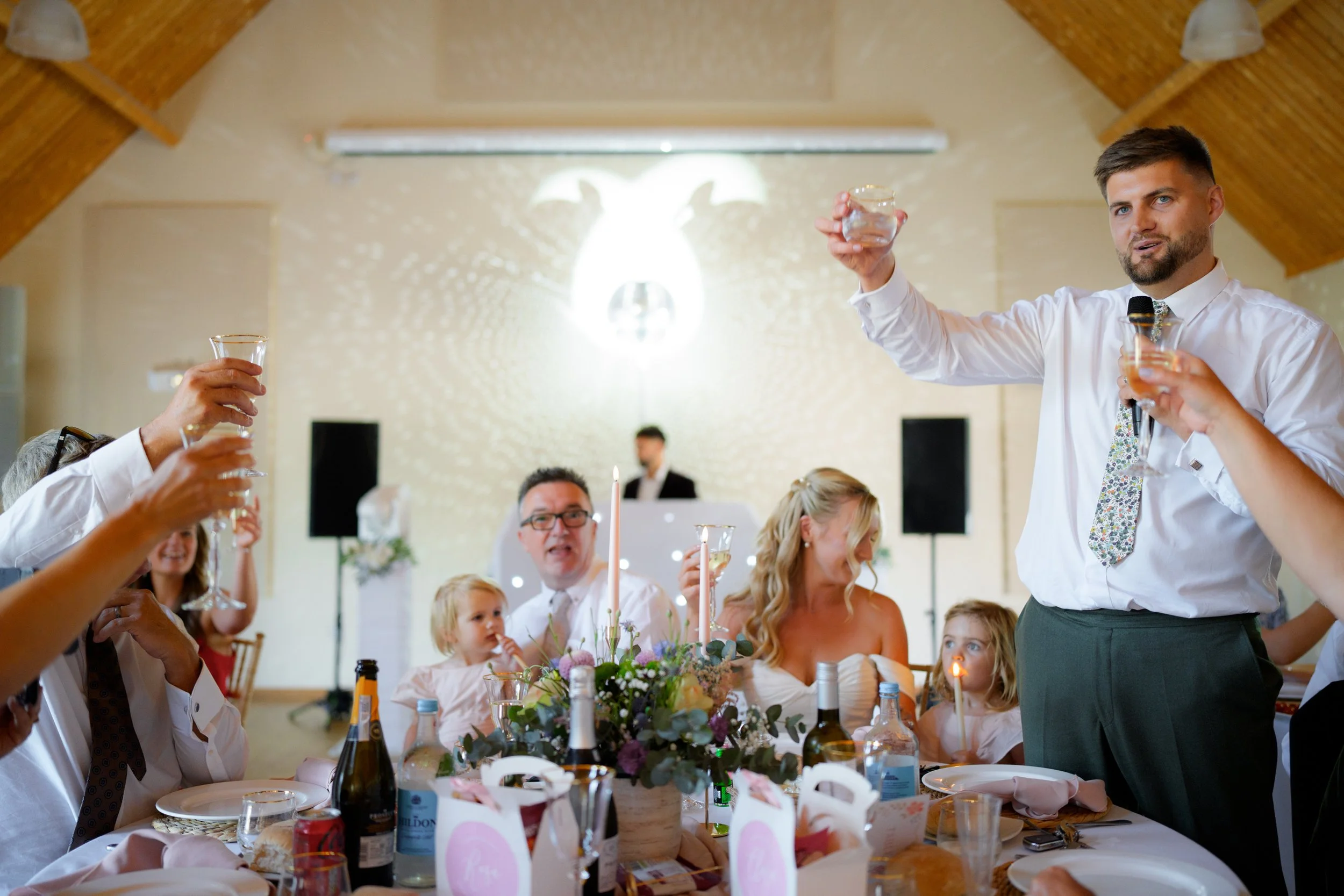 A man with a beard and short brown hair wearing a white shirt and patterned tie is giving a toast with a glass of champagne at a celebration dinner. He is surrounded by people of diverse ages sitting at a table with flowers, bottles, and candles in a