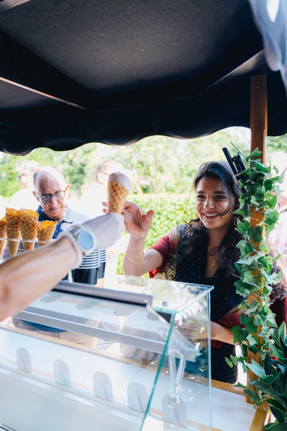 A woman in a colorful dress smiling as she receives an ice cream cone from a vendor at an outdoor ice cream stand with greenery in the background.