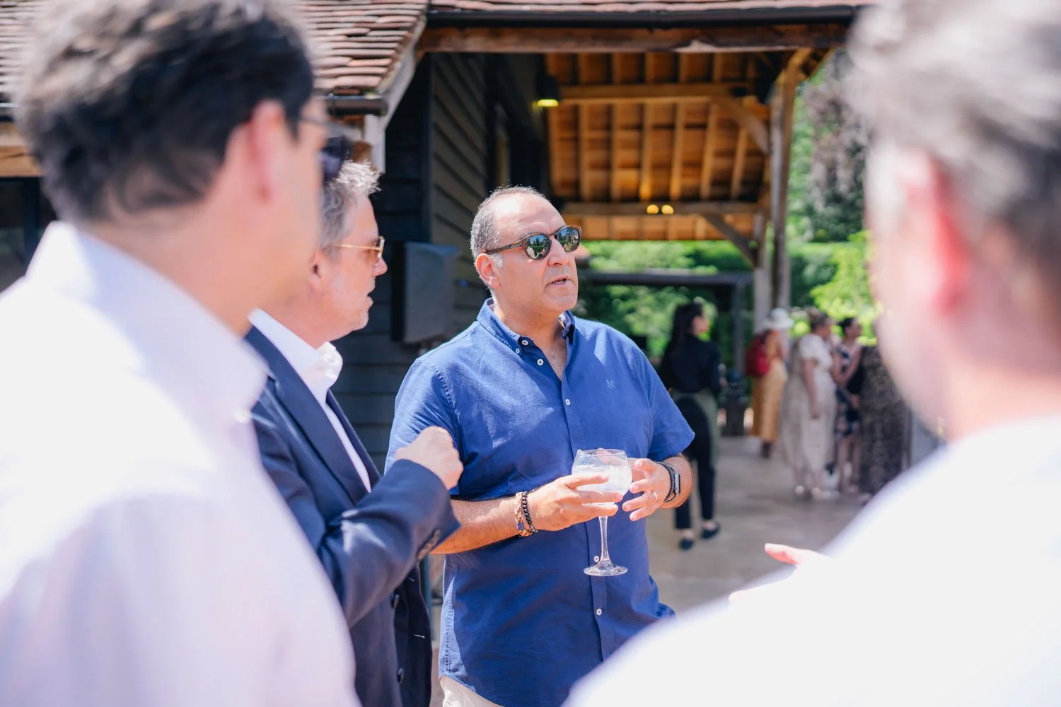 Man in blue shirt and sunglasses holding a wine glass, speaking to a group at an outdoor gathering.