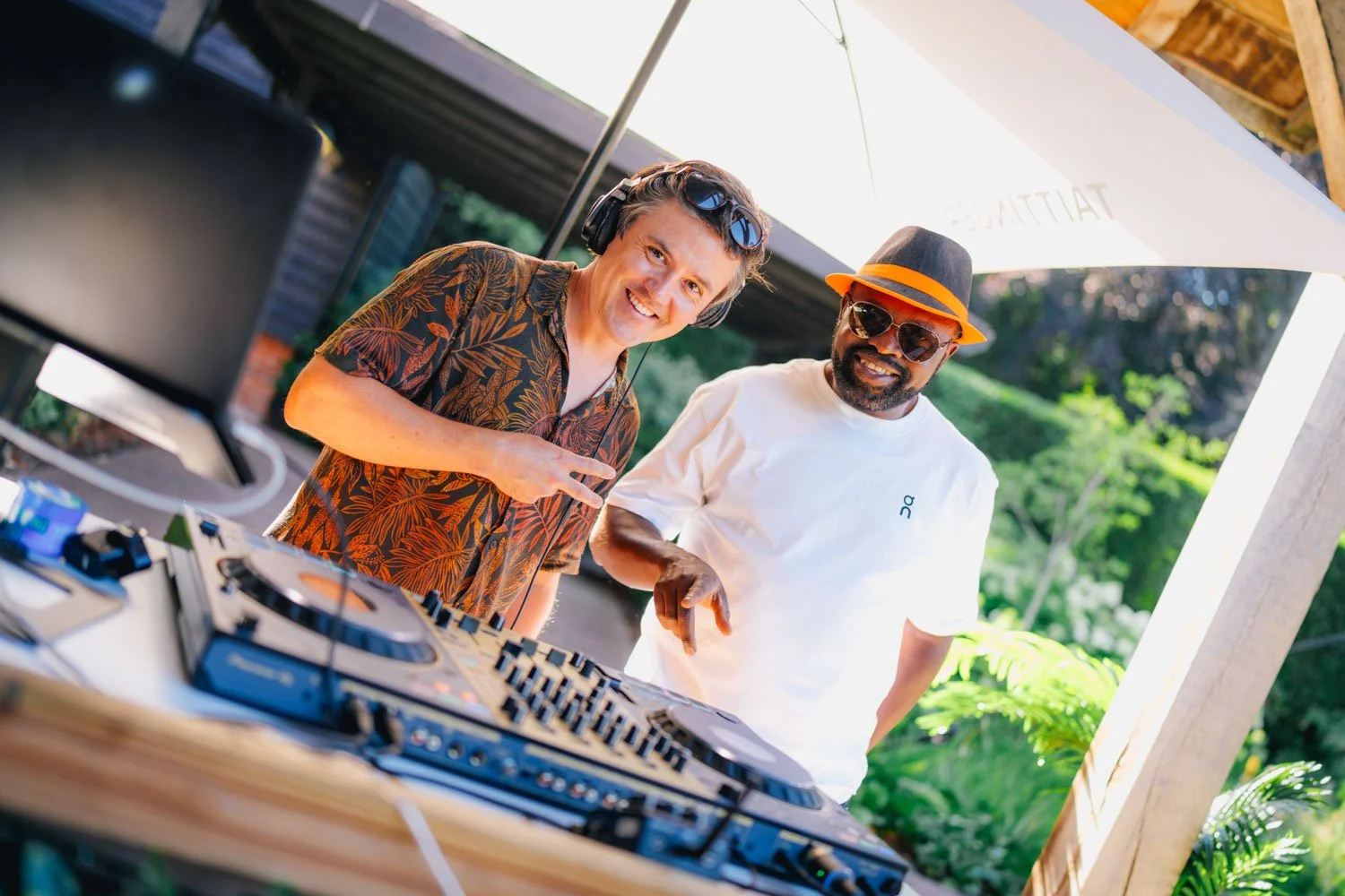 Two men, one with headphones and a tropical shirt and the other with sunglasses, a fedora, and a white t-shirt, standing at a DJ booth outdoors under a patio umbrella.