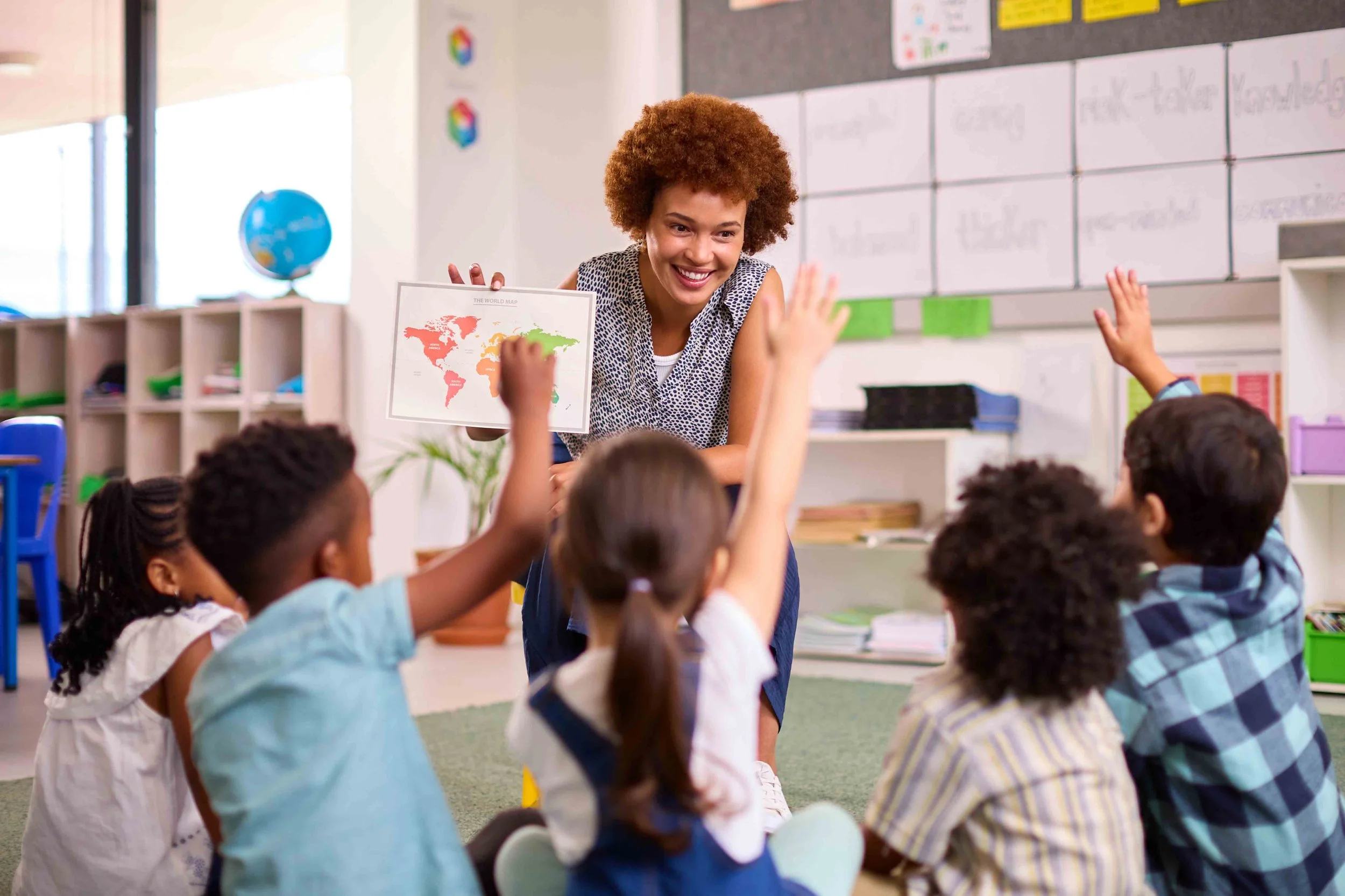 A classroom scene with a smiling female teacher with curly hair holding a world map while children raise their hands.