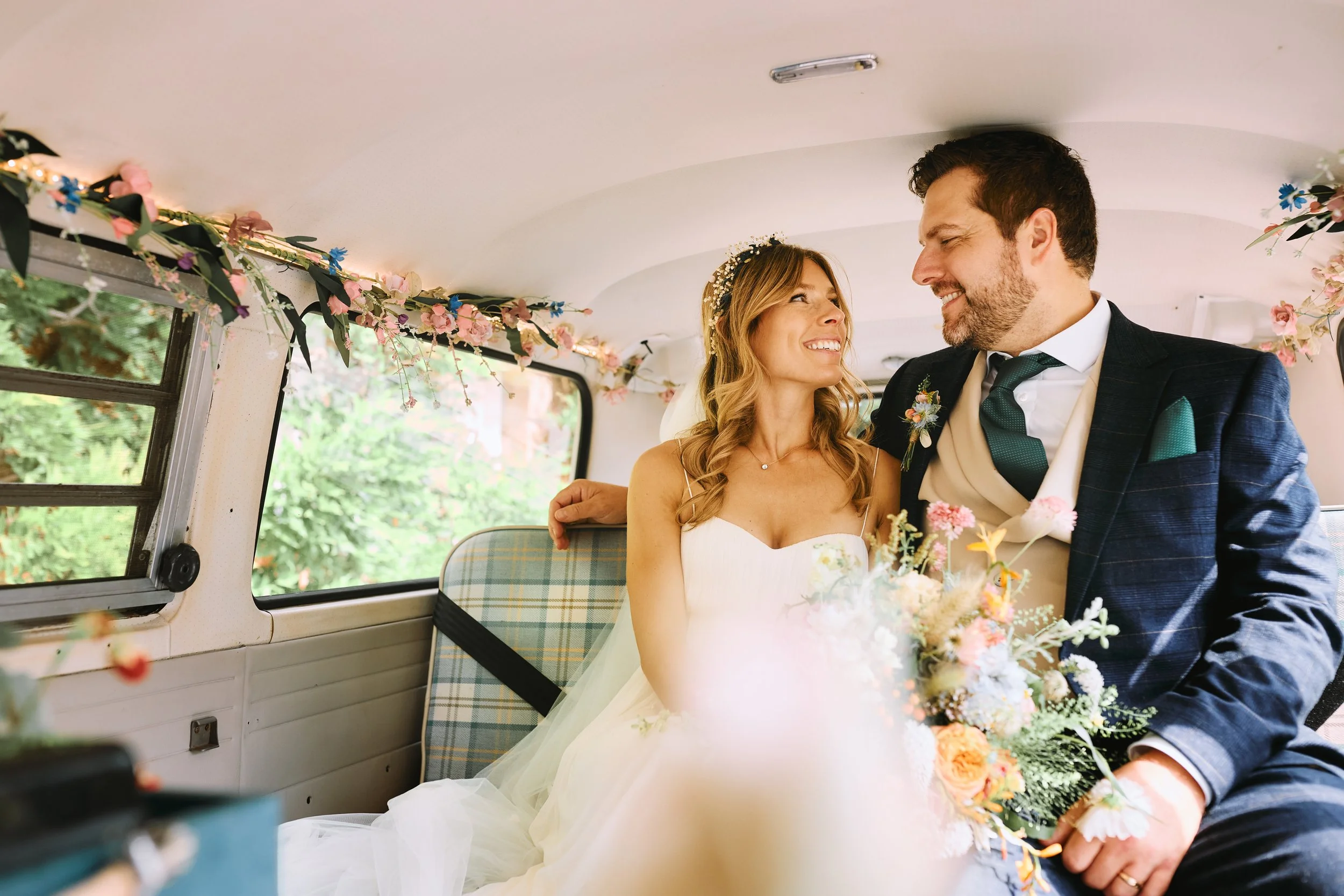 A bride and groom sitting together inside a decorated vehicle, smiling at each other, with floral decorations on the ceiling and a bouquet of flowers.
