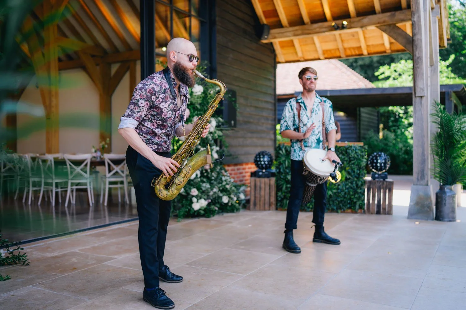 Two men playing musical instruments outdoors, one with a saxophone and the other with a drum, near a wooden building and greenery.