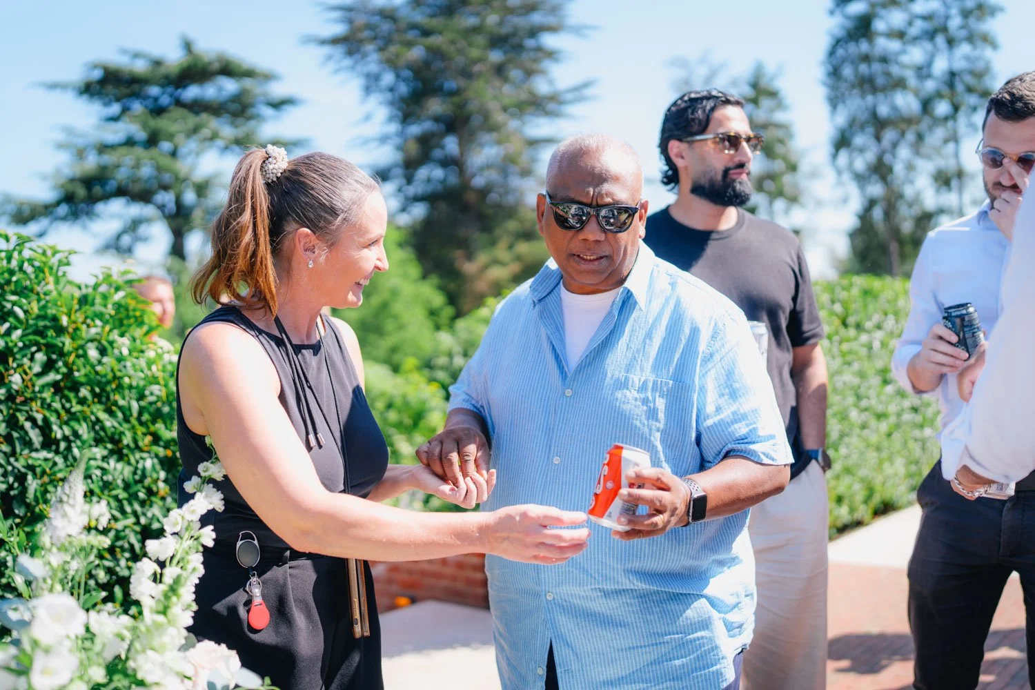 A group of people outdoors on a sunny day, engaging in conversation with greenery in the background. A woman is smiling and showing something to a man wearing sunglasses, who is holding a can.
