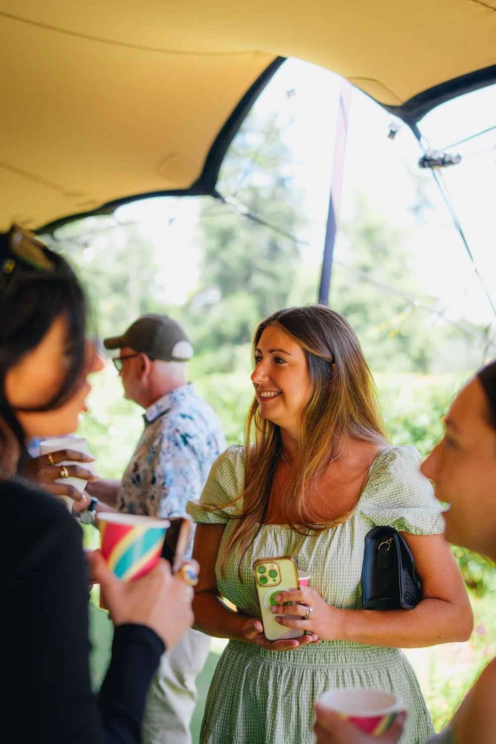 A group of people enjoying a sunny outdoor gathering, with a woman in a light green dress smiling and holding a phone, surrounded by others with cups, under a tent.