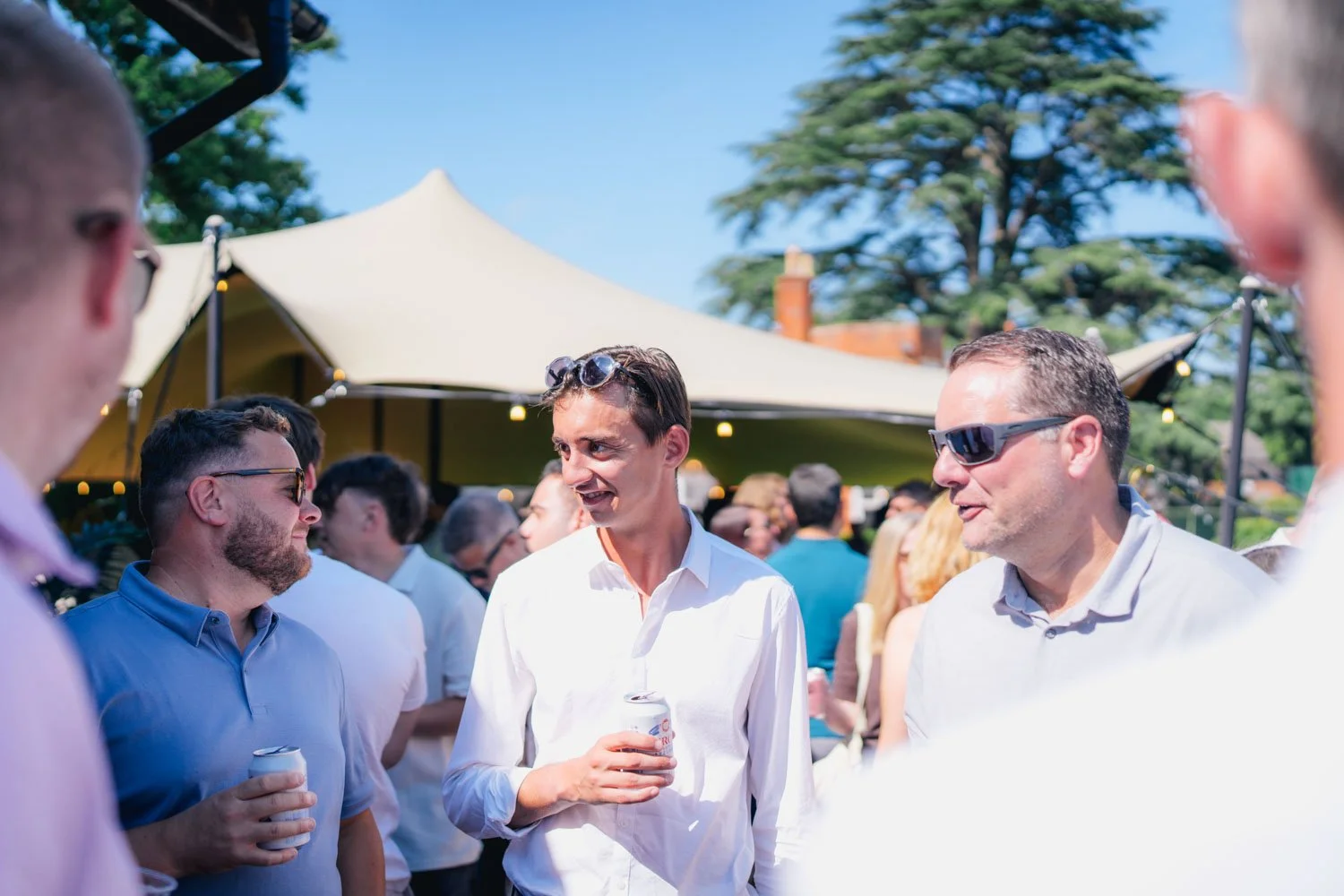 Group of men conversing at outdoor event on sunny day, with canopy tent and trees in background.