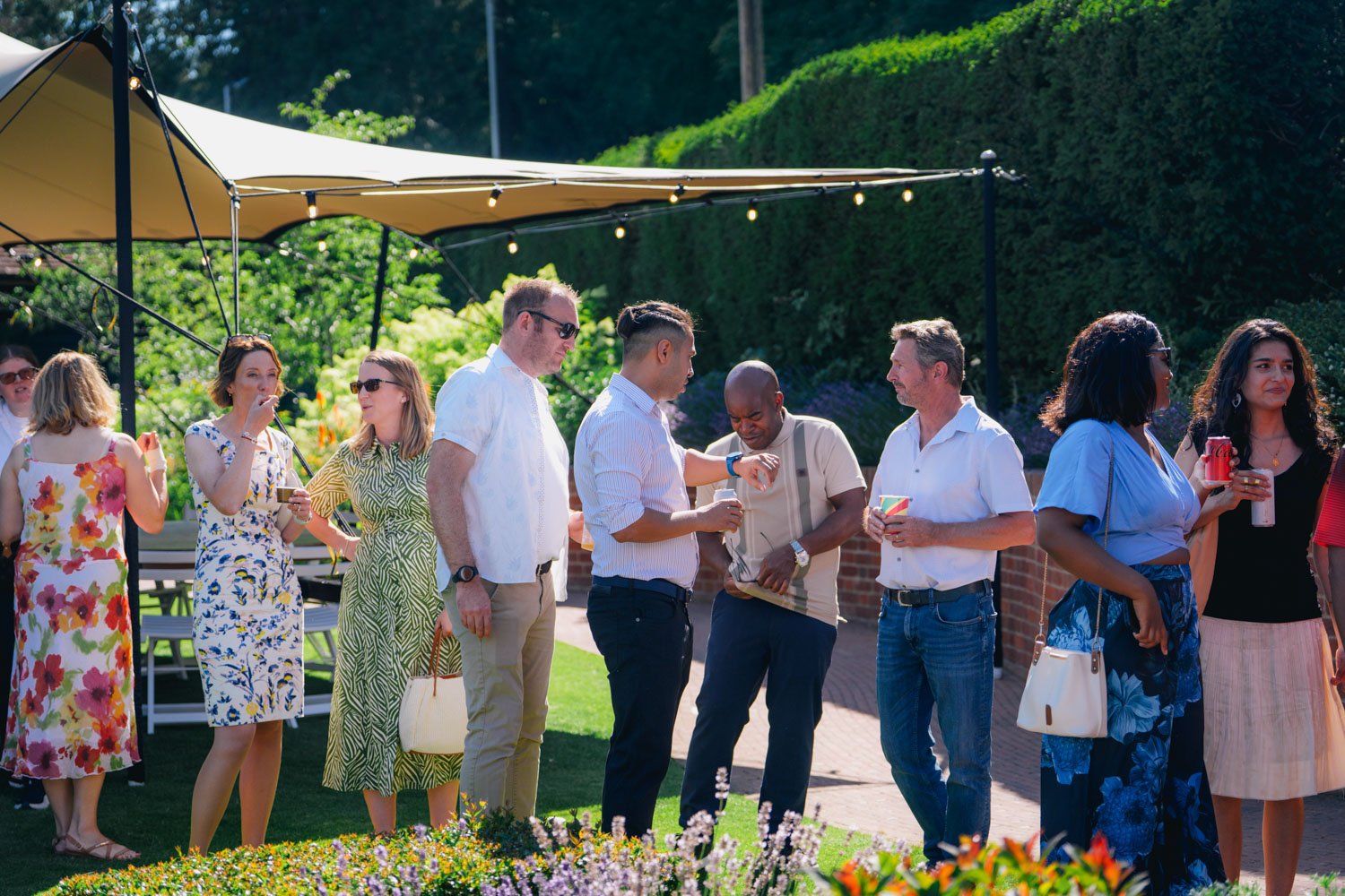 A group of people at an outdoor gathering under a beige canopy with string lights, socializing and holding drinks, with green bushes and flowers in the background on a sunny day.
