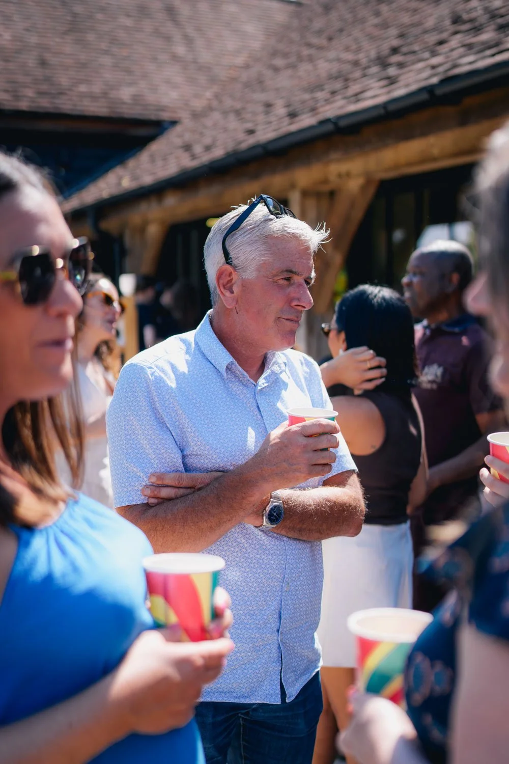 A group of people socializing outdoors at a sunny gathering, holding colorful cups, with a rustic building in the background.