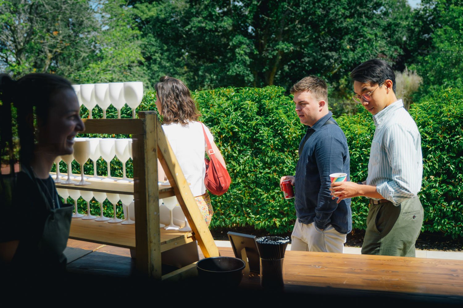 People standing in line outdoors, holding cups and cans, with a display of glassware and greenery in the background.