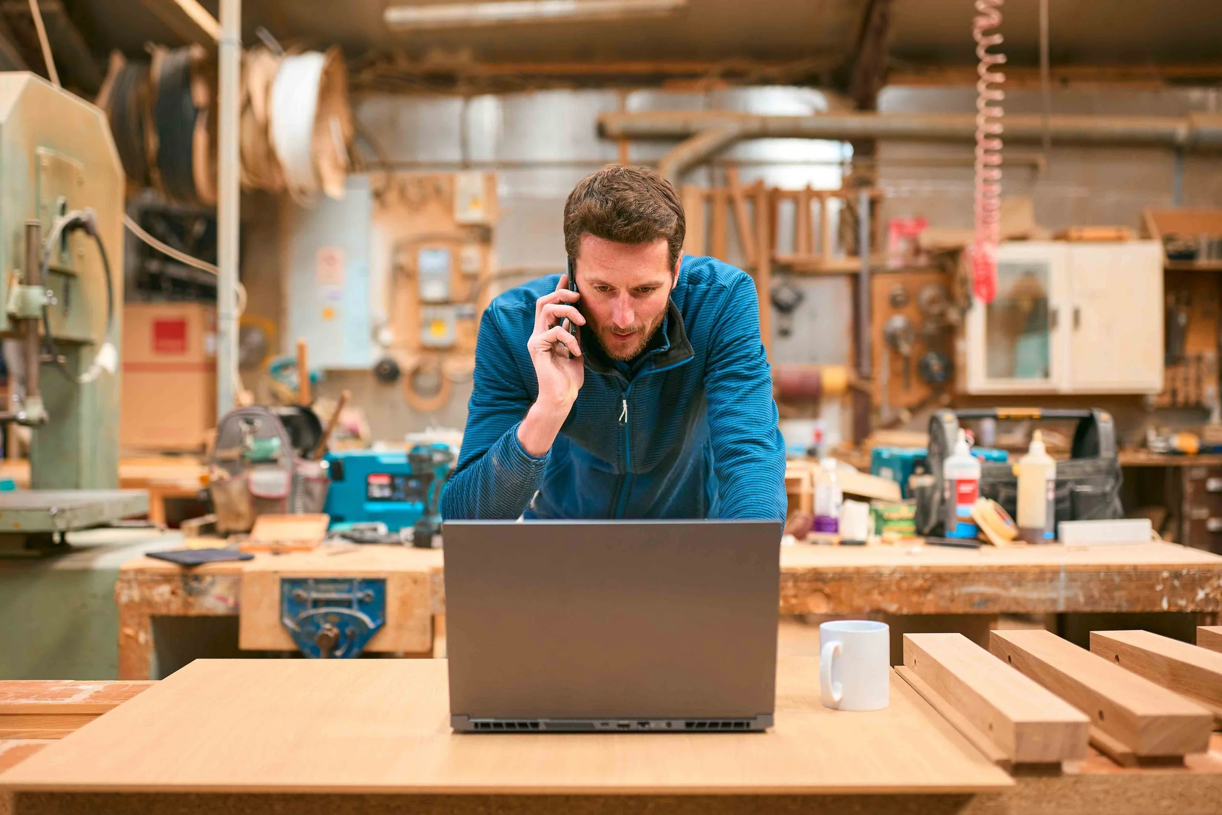 A man is talking on a cellphone while working in a woodworking shop with tools and woodwork materials around him.