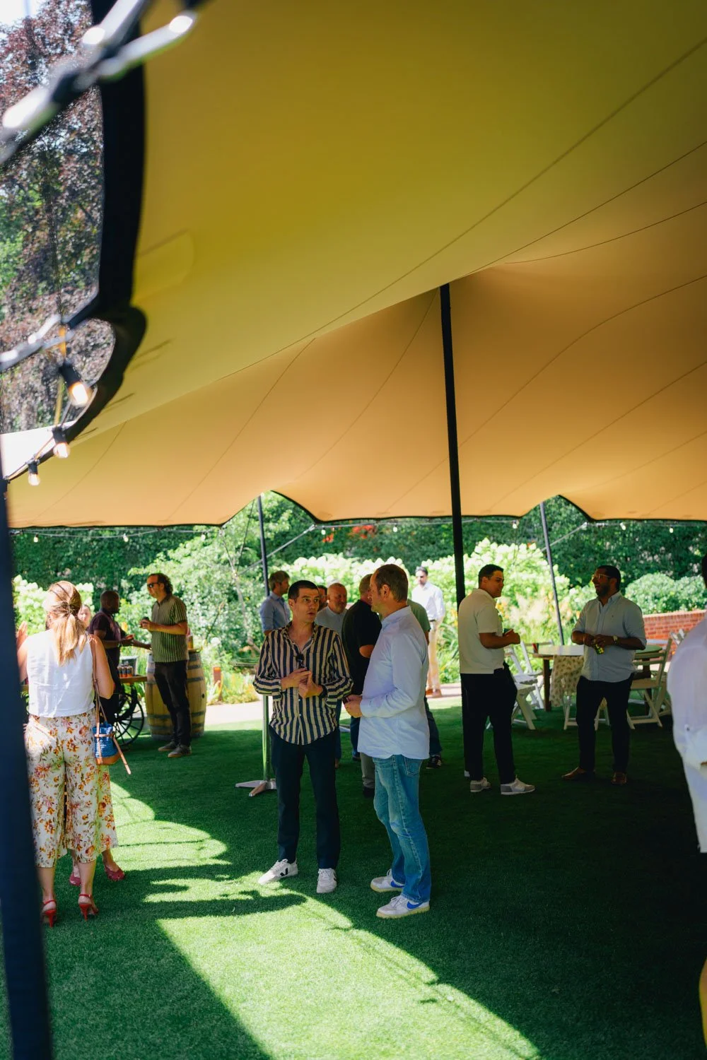 People socializing under a large yellow canopy in an outdoor garden setting.