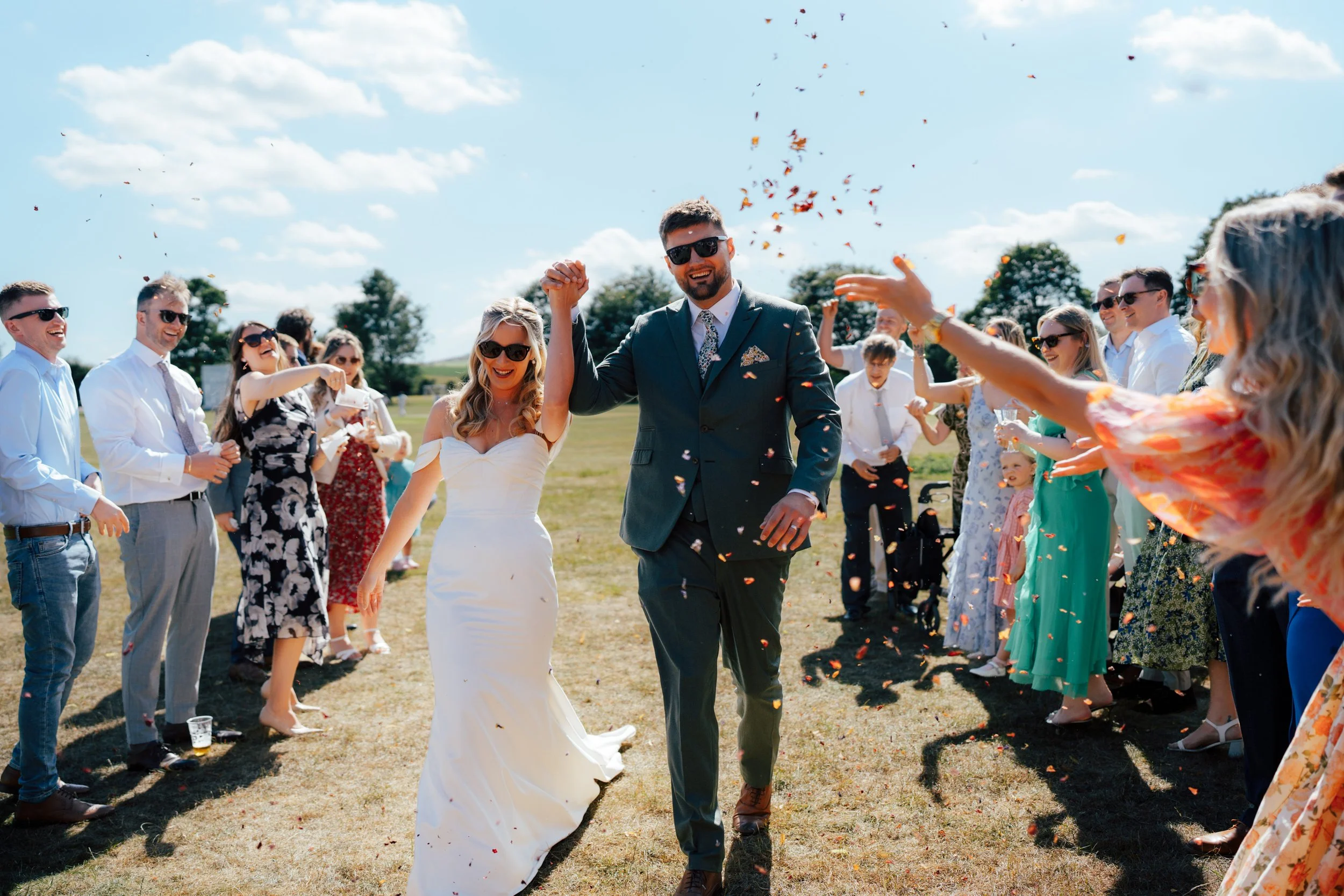 A bride and groom walking hand in hand through a confetti shower during their outdoor wedding celebration, surrounded by smiling guests wearing sunglasses and summer dresses on a sunny day.