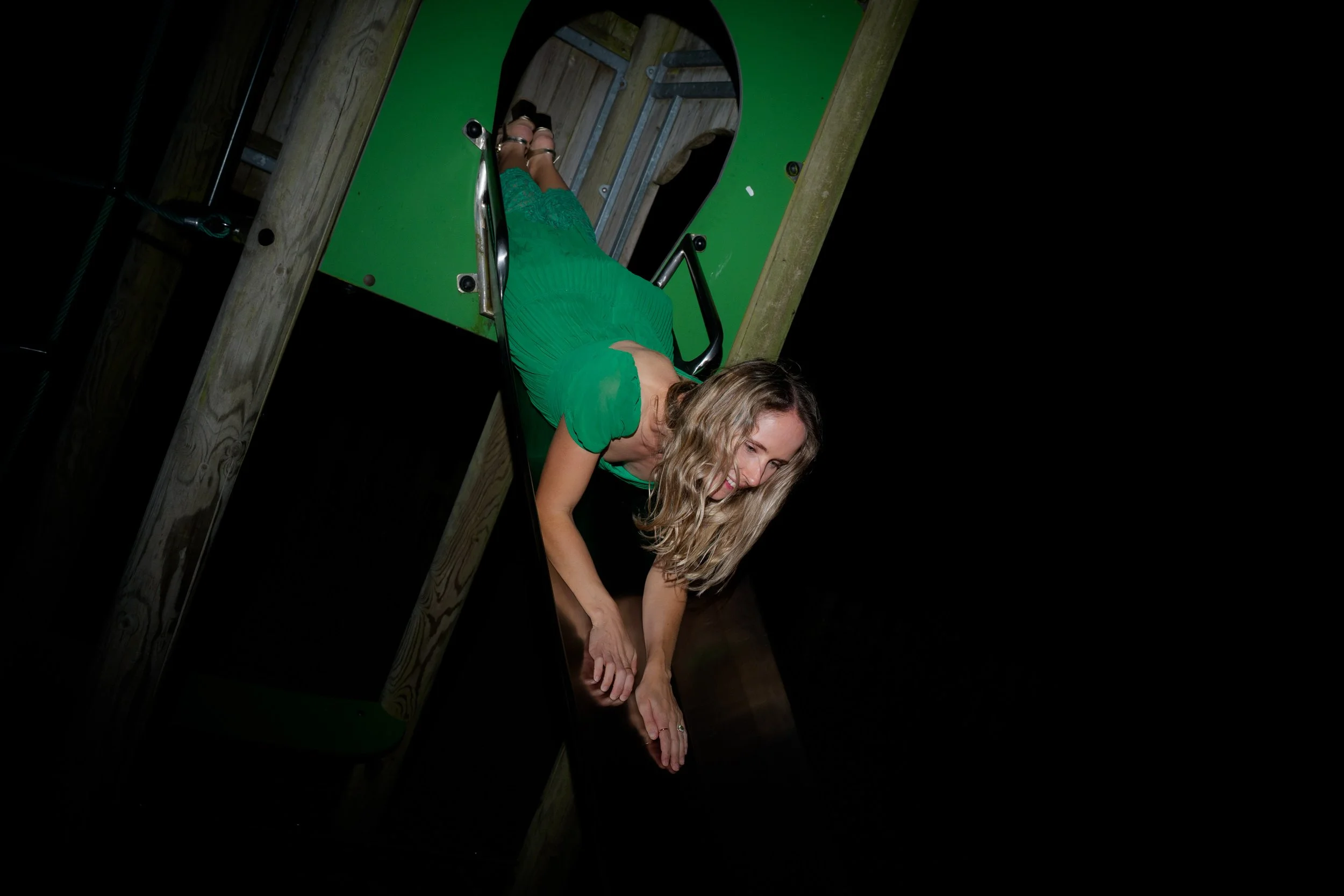 Woman in a green dress hanging upside down on a playground slide, holding her ankles.